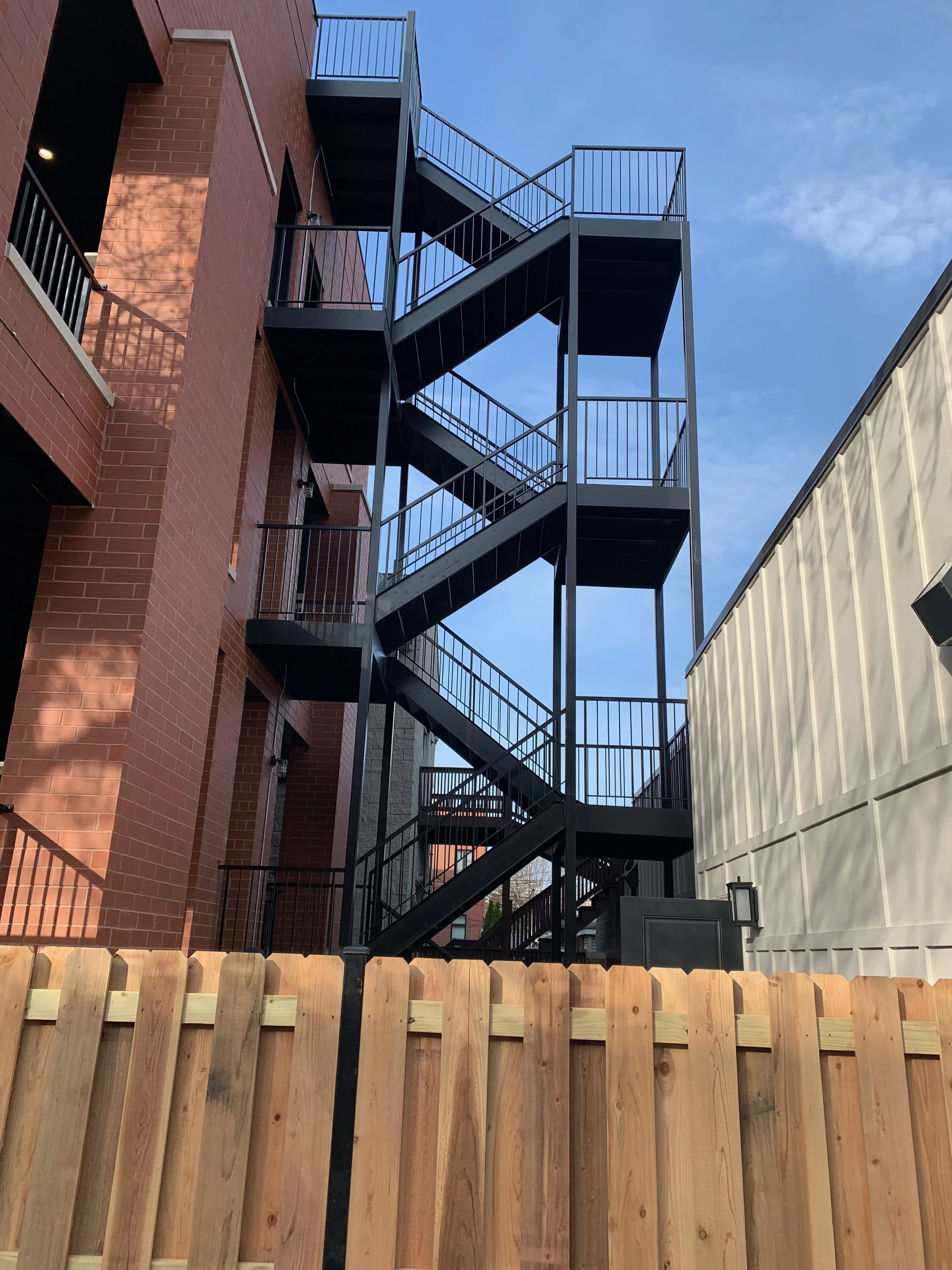 Black metal fire escape staircase attached to a red brick building, with a light-colored wooden fence in the foreground and a clear blue sky overhead.
