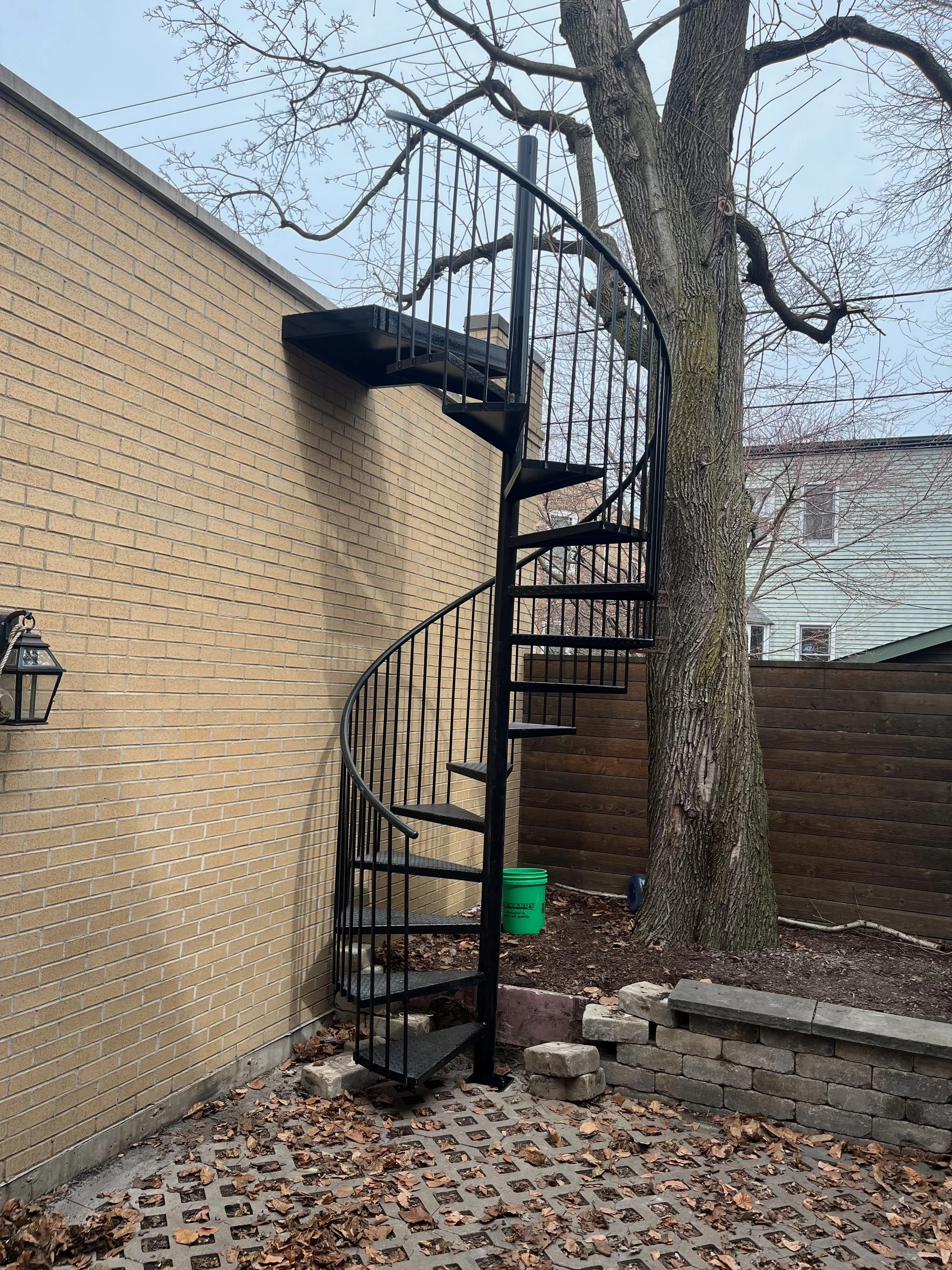 Black metal spiral staircase outside next to a beige brick wall, with a tree and fallen leaves on the ground.