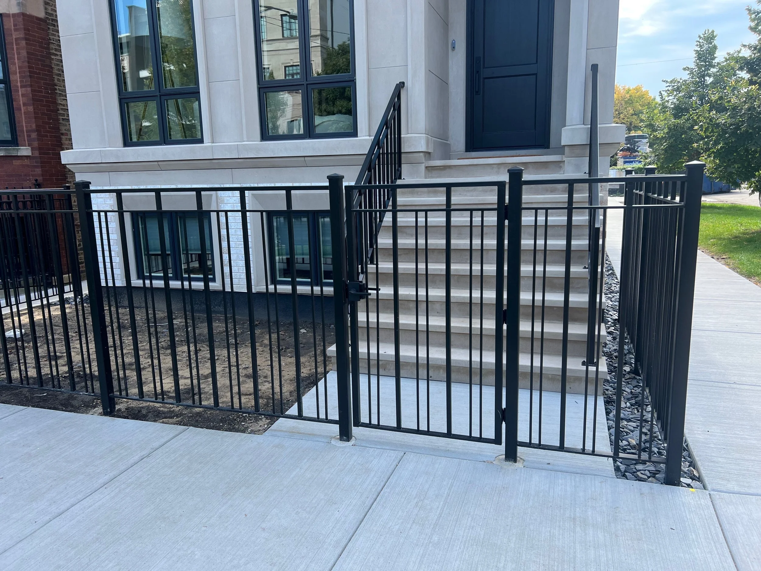 Exterior view of modern townhouse with black front door, large windows, stairs leading to the door, an underground basement with windows, and a black metal fence surrounding the property.