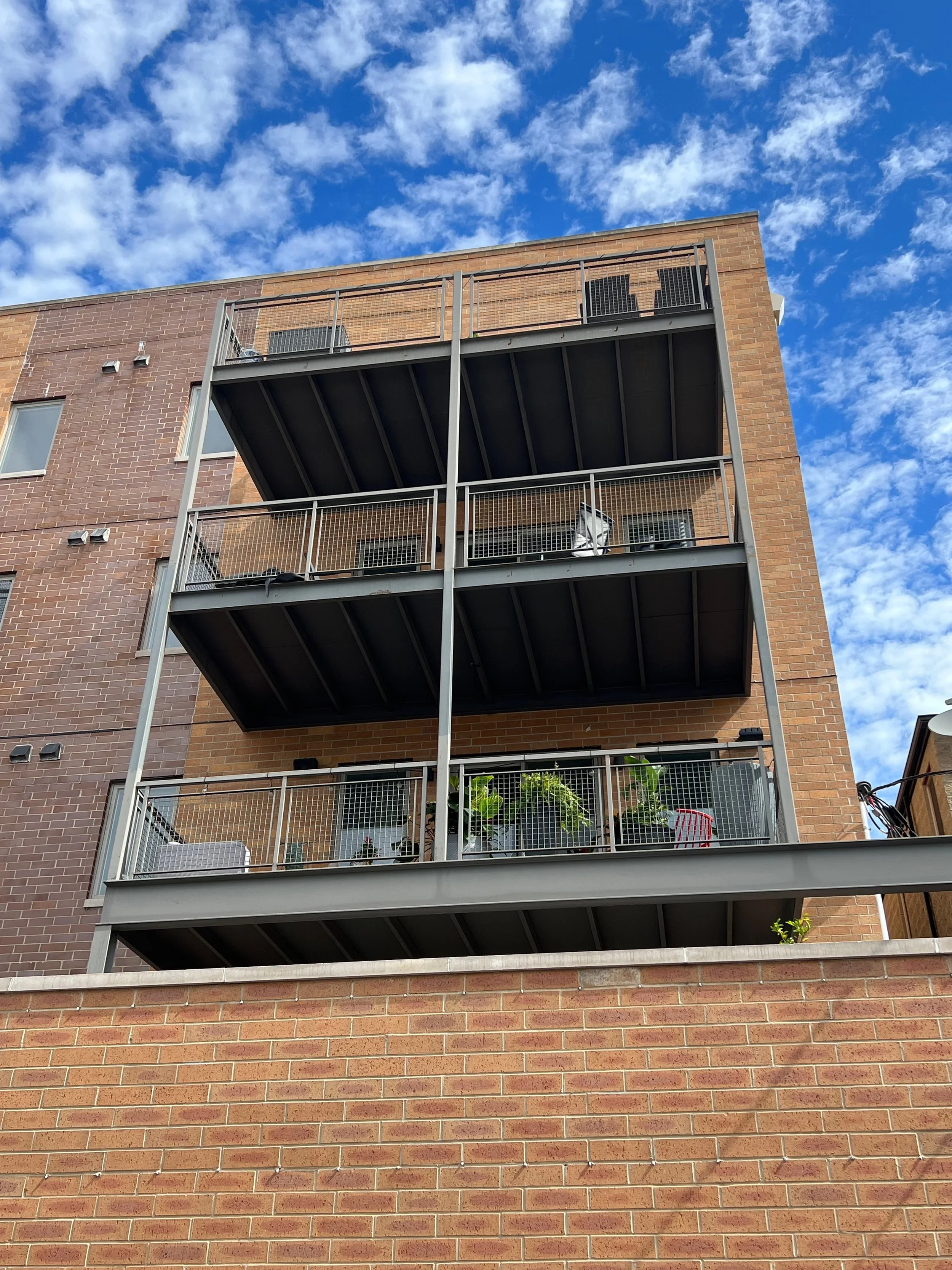 A brick apartment building with three floors visible, each with a balcony that has plants, furniture, and items. The sky above is bright blue with scattered white clouds.