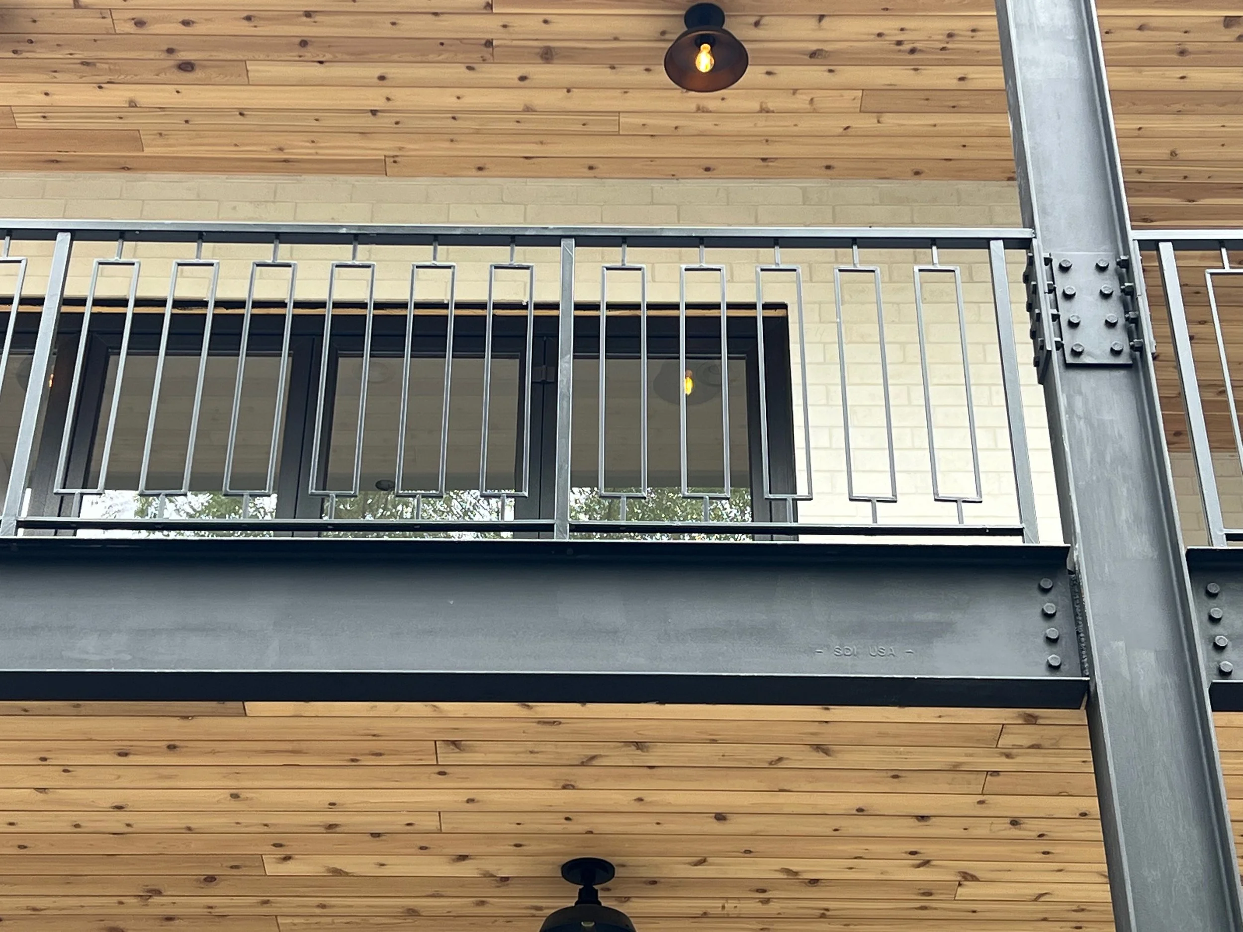 A balcony with metal railing in front of a yellow brick wall and a wooden ceiling with a hanging light bulb.