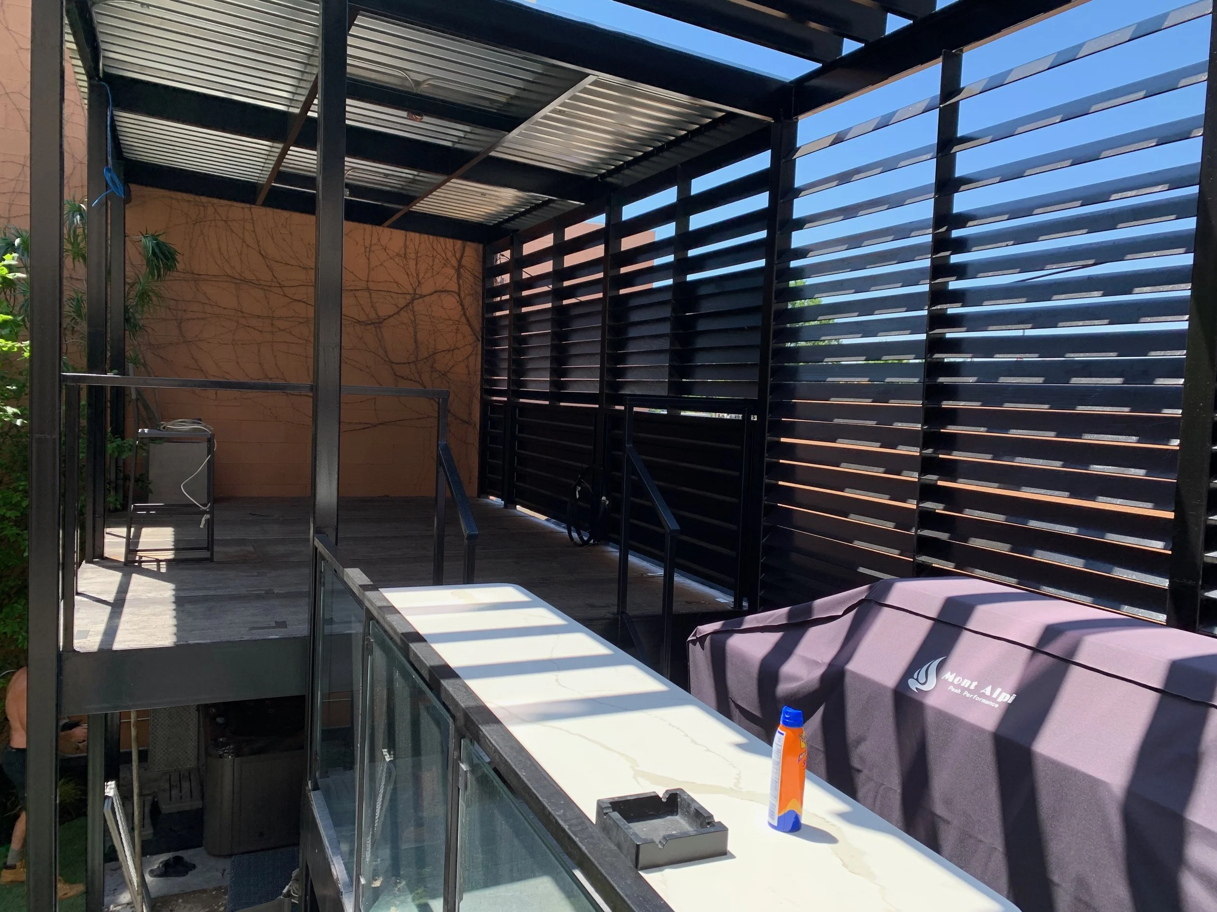 Construction site with a metal framework and slatted black privacy screens, a gray countertop with building tools and supplies, and a beige wall with a vine-like pattern in the background.