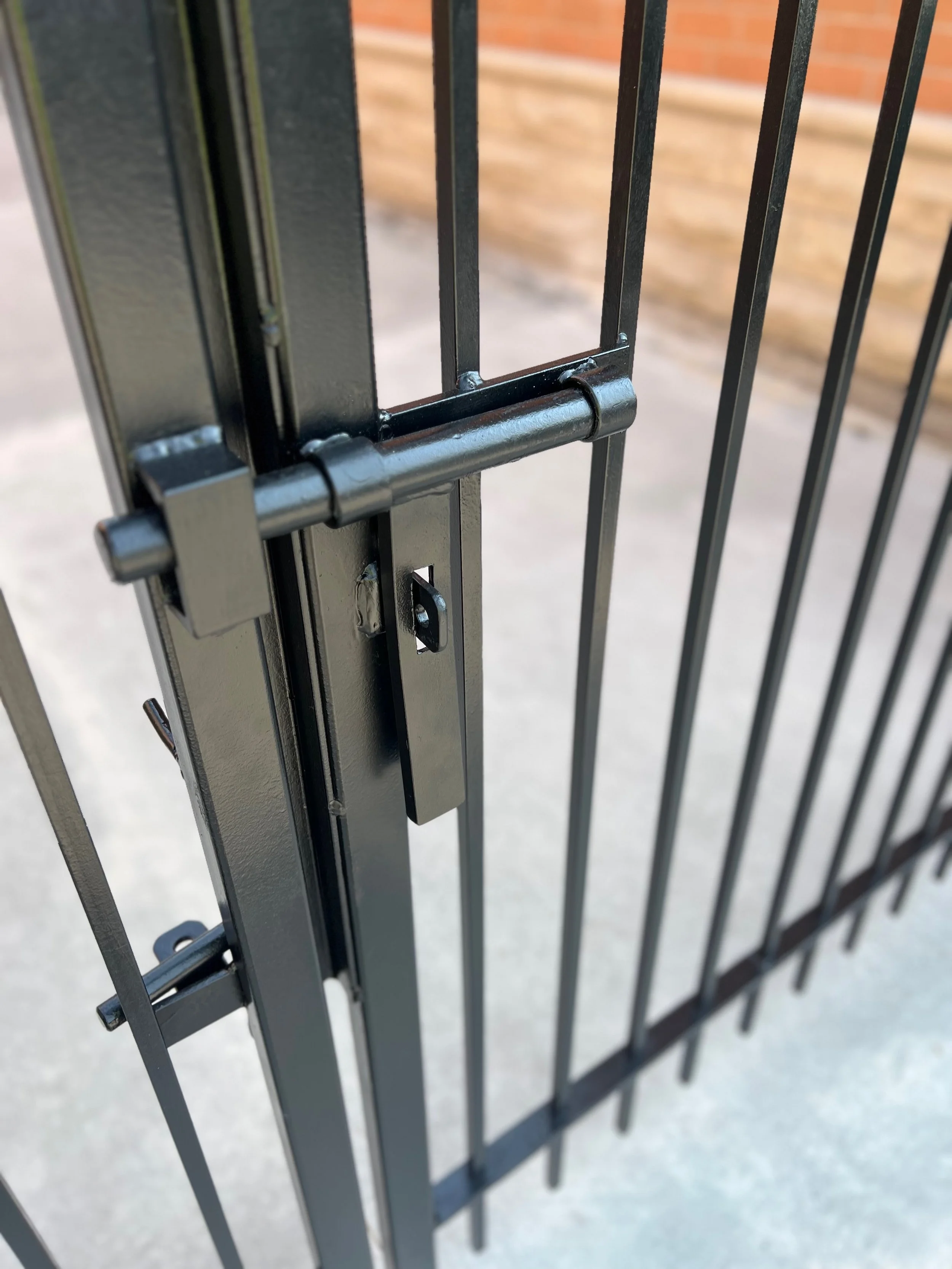 Close-up of a black metal gate with a latch, with a brick wall in the background.
