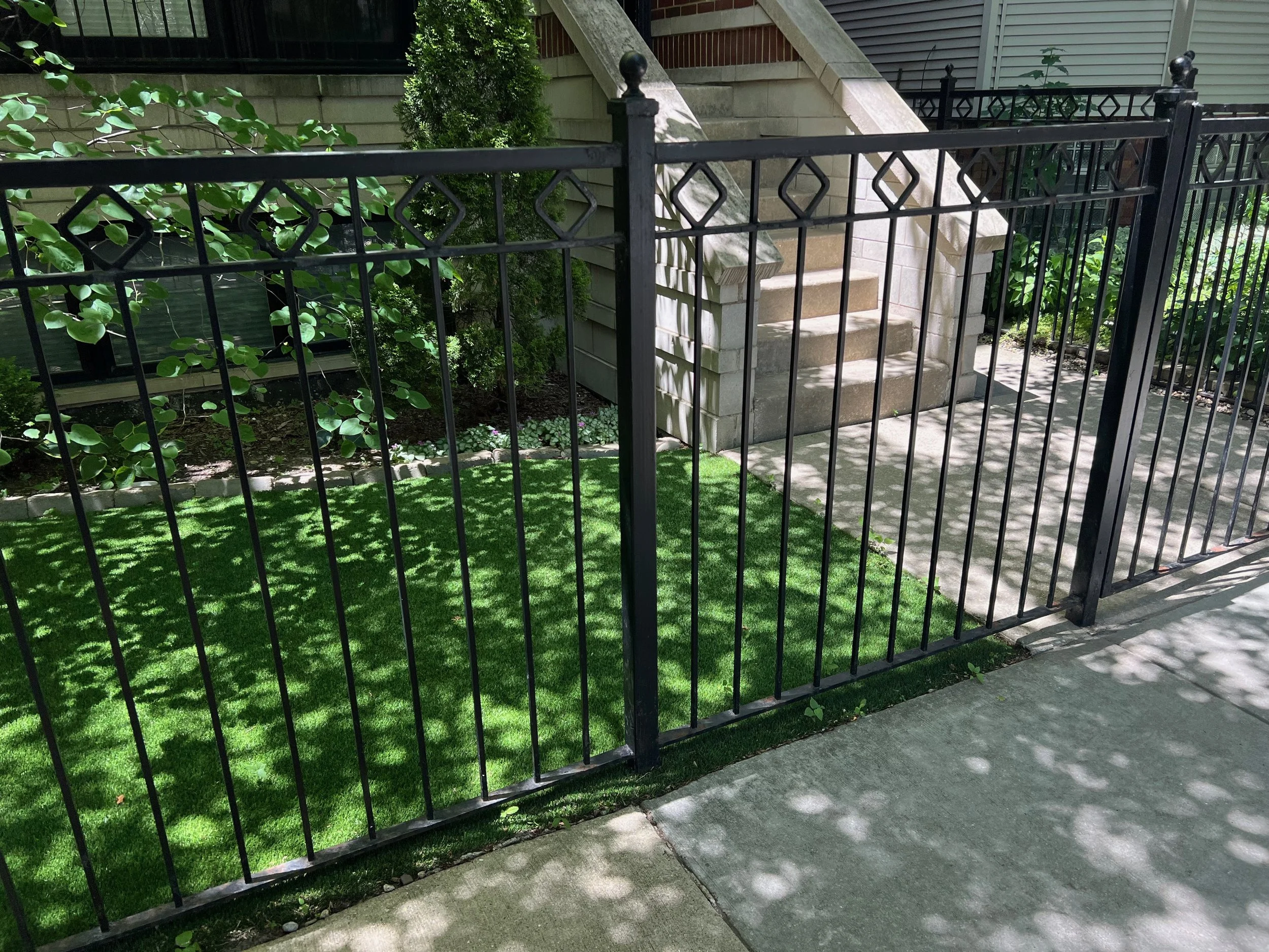 Black metal fence surrounding a small landscaped area with green grass, bushes, and a concrete staircase leading to a building.