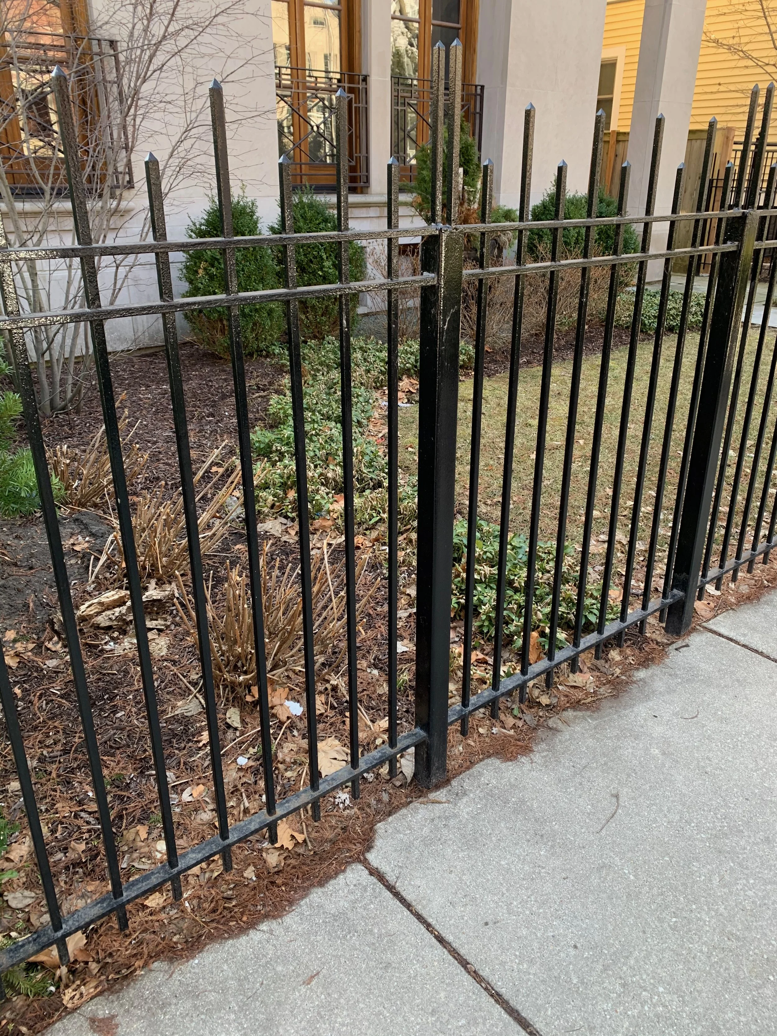 A black metal fence surrounds a small garden bed with leafless and green bushes, situated in front of a beige and yellow building with windows and railings.