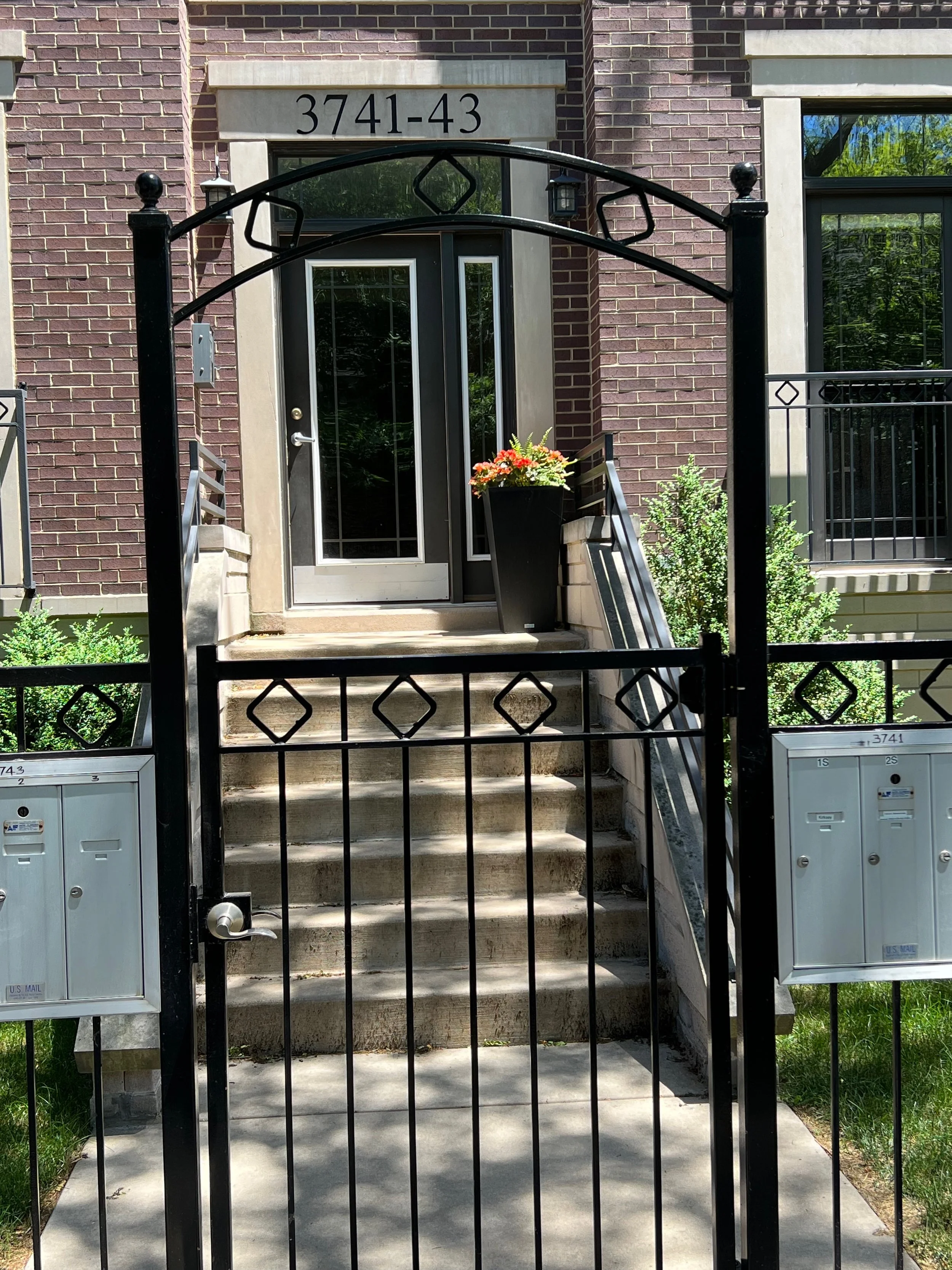 Front entrance of a residential building with stairs leading up to a door, a black metal gate, and mailboxes on each side of the stairs, with a flower pot on the right side of the door.
