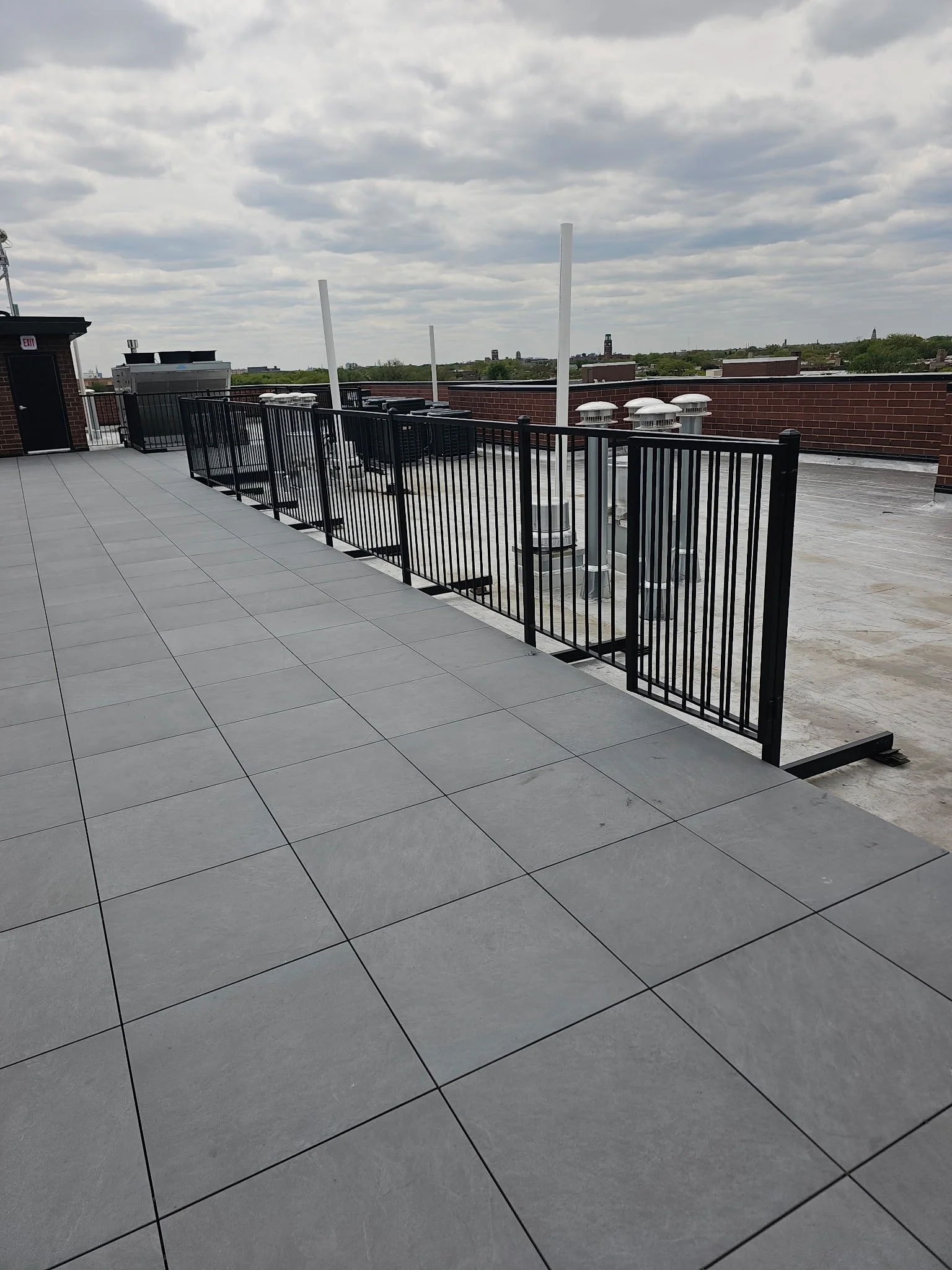 Rooftop view with gray tiled floor, black metal railing, and industrial HVAC units against a cloudy sky.