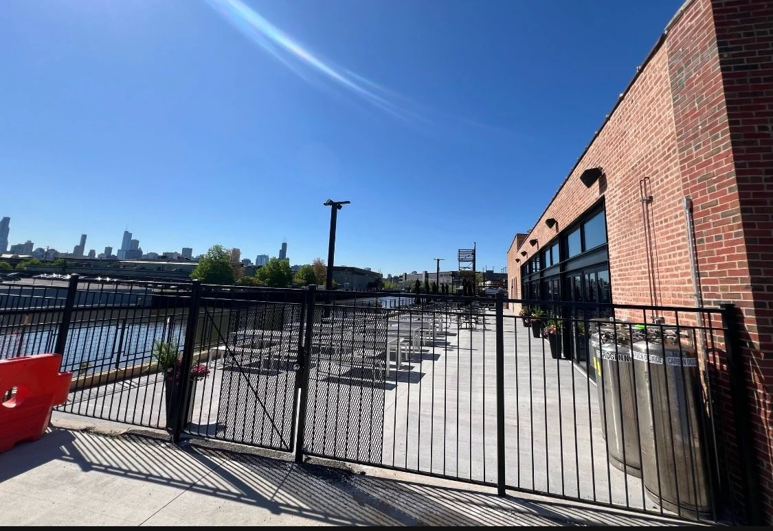 Rooftop patio with black metal fence, potted plants, and city skyline in the distance against a clear blue sky.