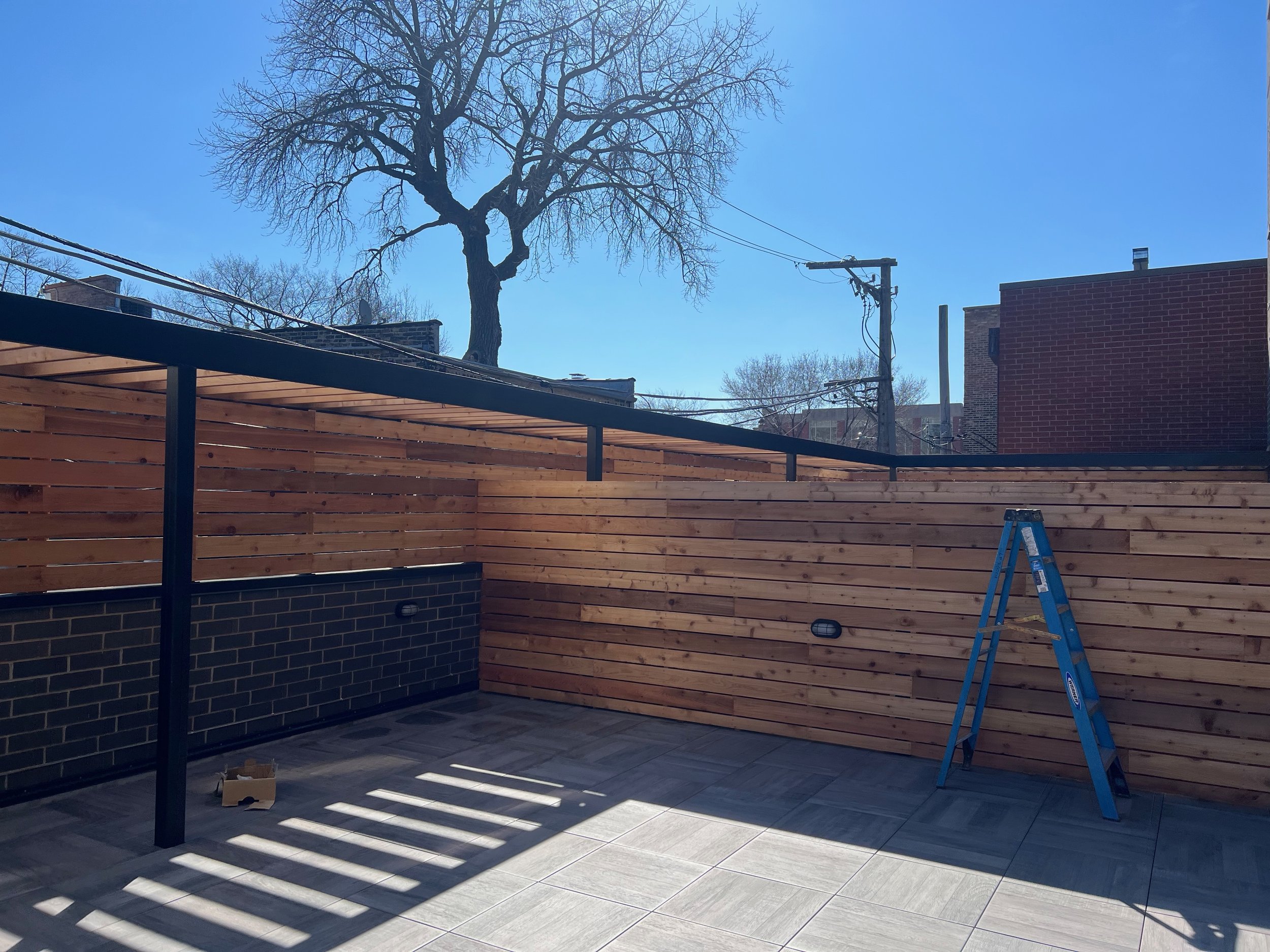 Construction site with a wooden fence, a tree, a ladder, and neighboring buildings on a sunny day.