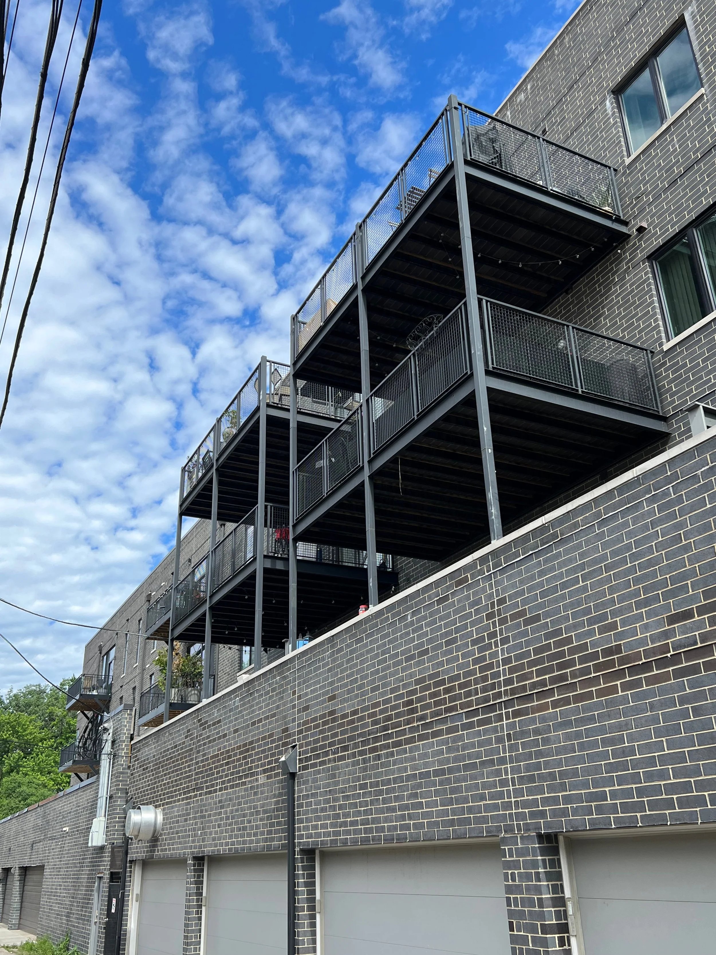Multiple black metal fire escape balconies attached to a dark brick residential building under a partly cloudy blue sky.