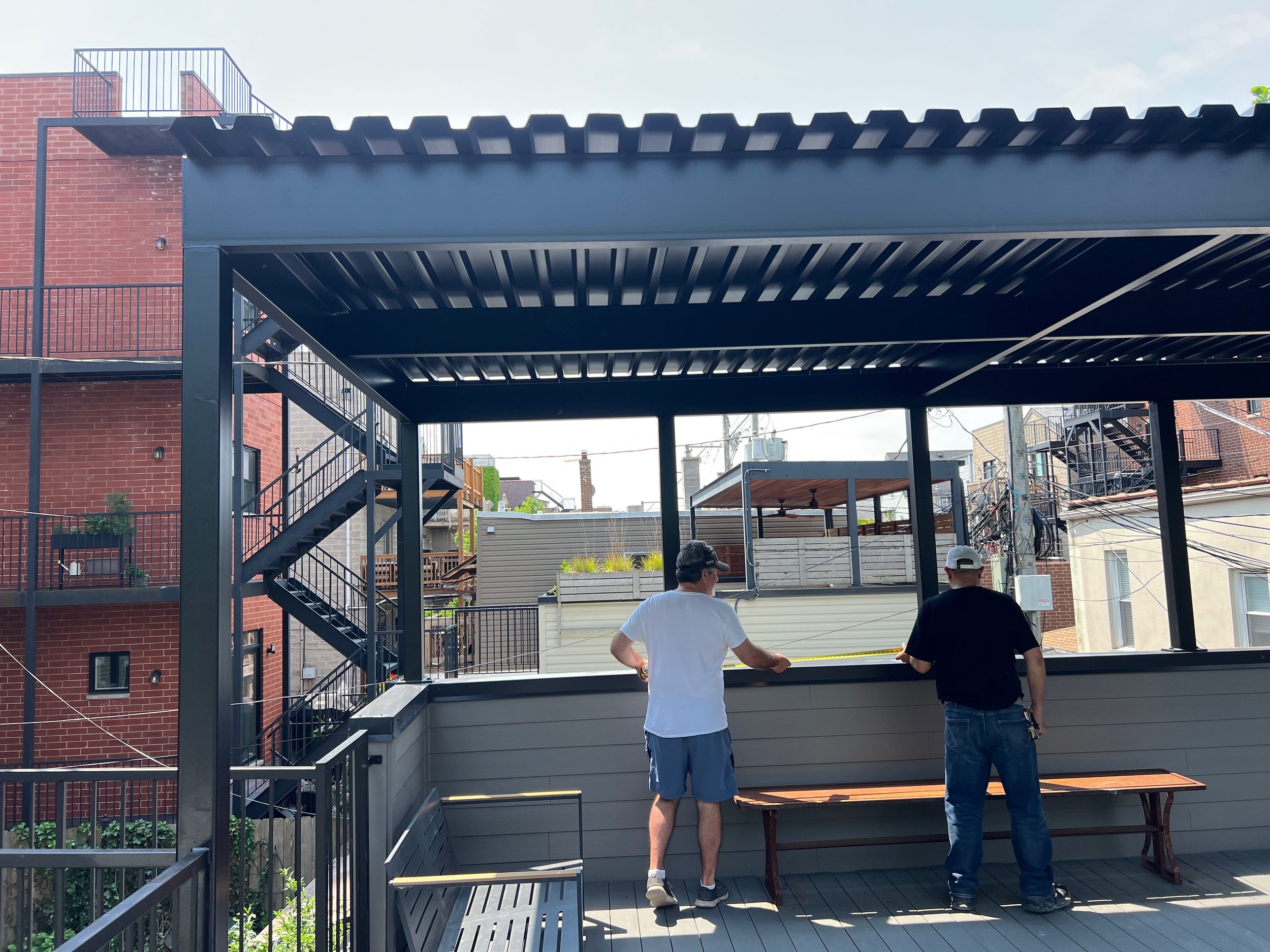 Two men standing on a deck, viewing a newly constructed modern outdoor patio with metal-framed structures, stairs, and landscaping, in an urban residential area.