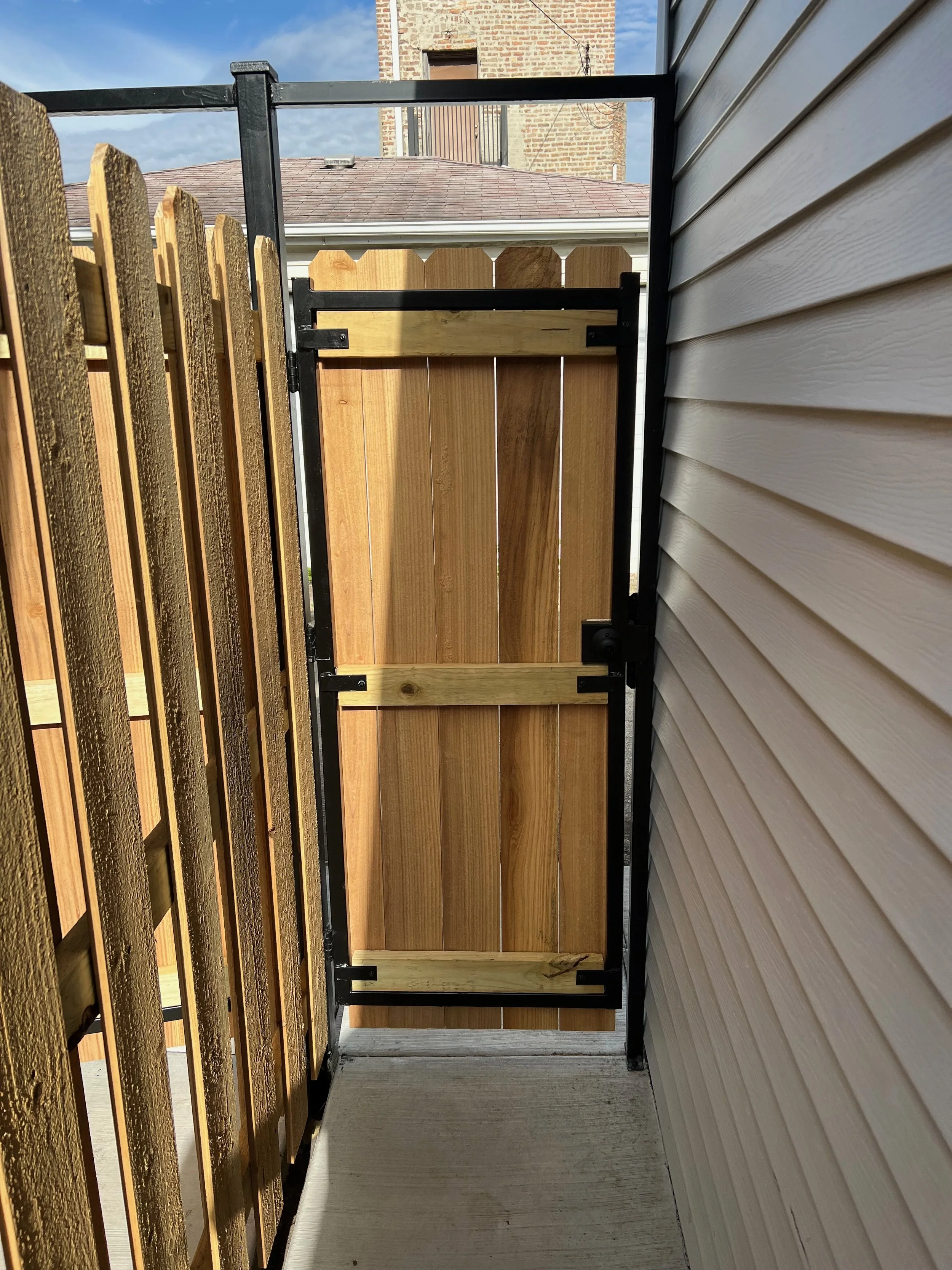 View from a small balcony or porch with a black metal railing and wooden privacy screen, beige siding on the right, neighboring brick building with a rooftop and sky in the background.