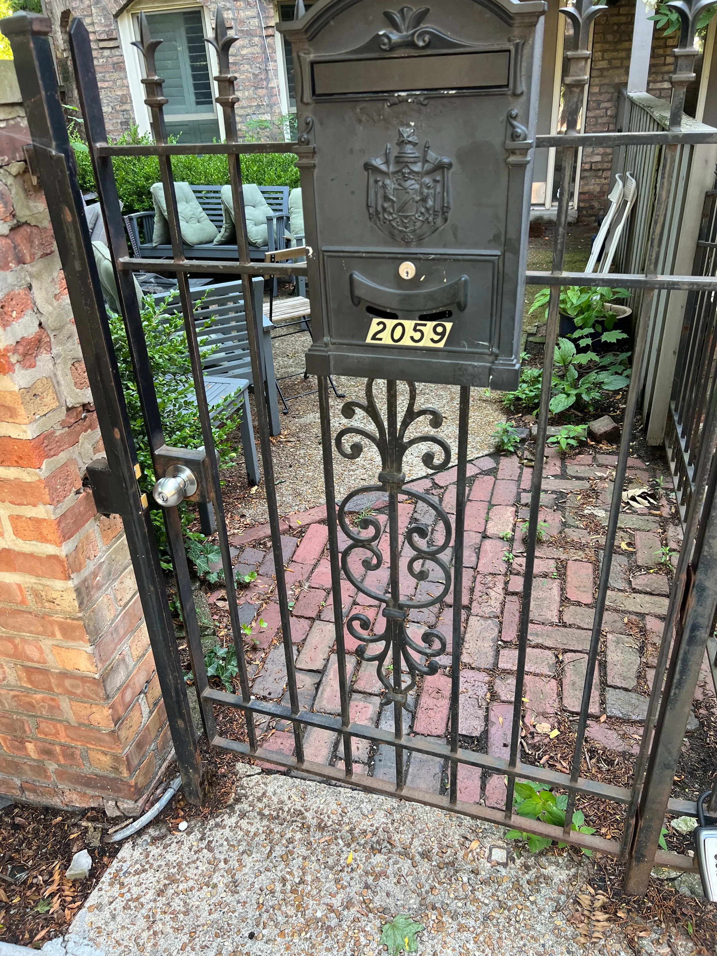 Black wrought iron gate with a mailbox labeled 2059 in front of a brick house with patio furniture and greenery in the background.