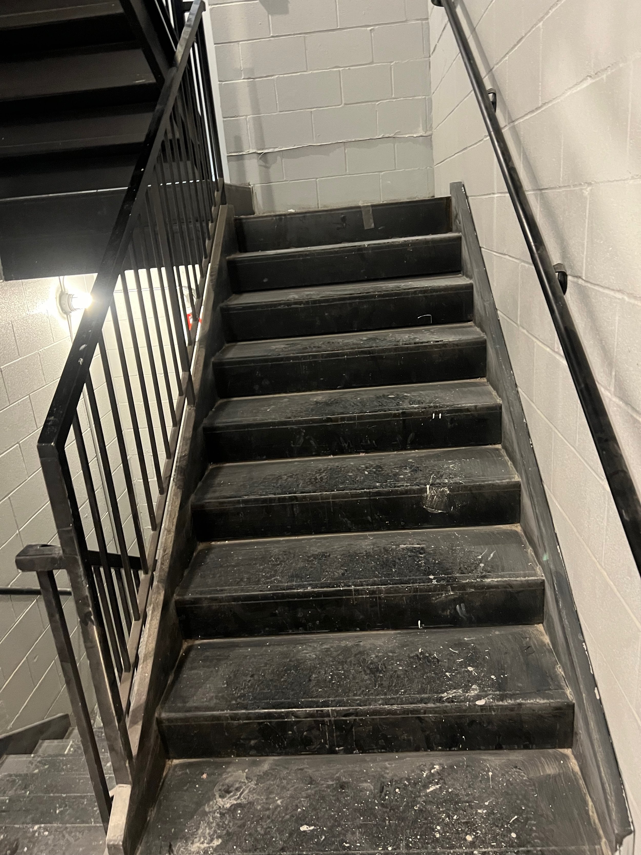 Black staircase with a metal handrail on the right side, against gray brick wall, with visible dirt and debris on the steps.