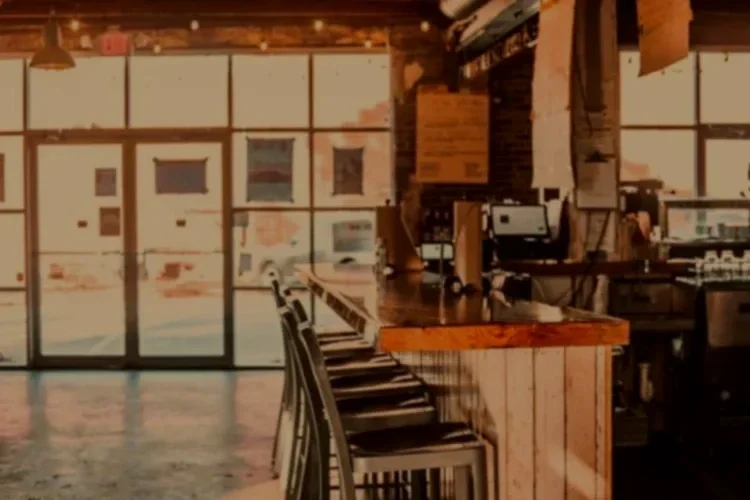 Interior of a rustic coffee shop or cafe with wooden accents, high tables, and bar stools near large glass doors, sunlight streaming in.