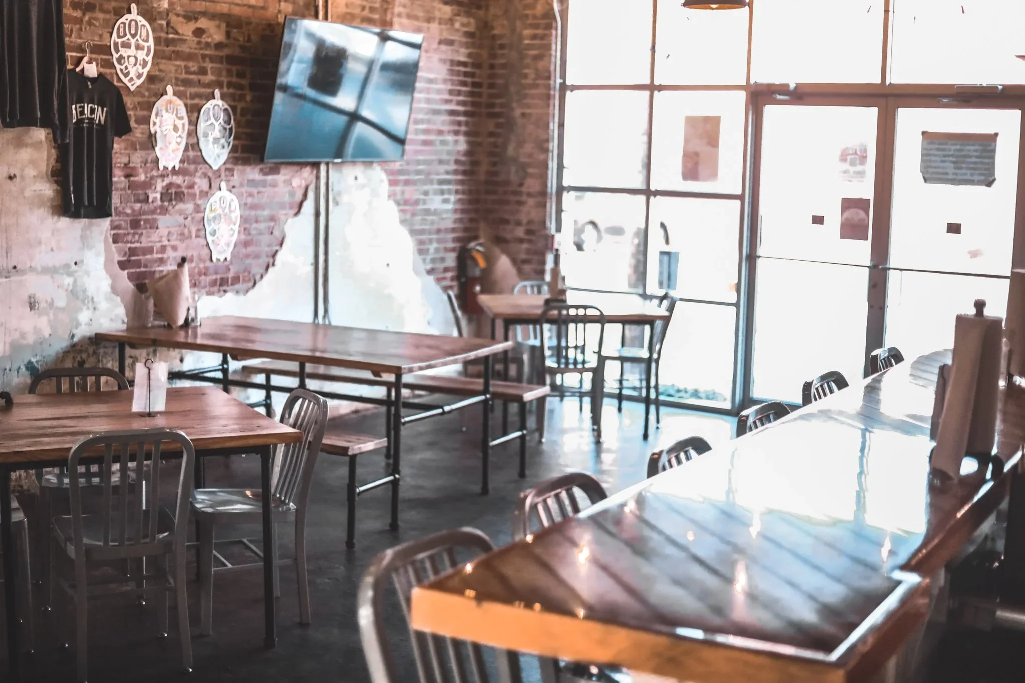 Empty restaurant or cafe interior with brick walls, wooden tables, metal chairs, large windows, a television mounted on the wall, and some art or decorations on the brick wall.