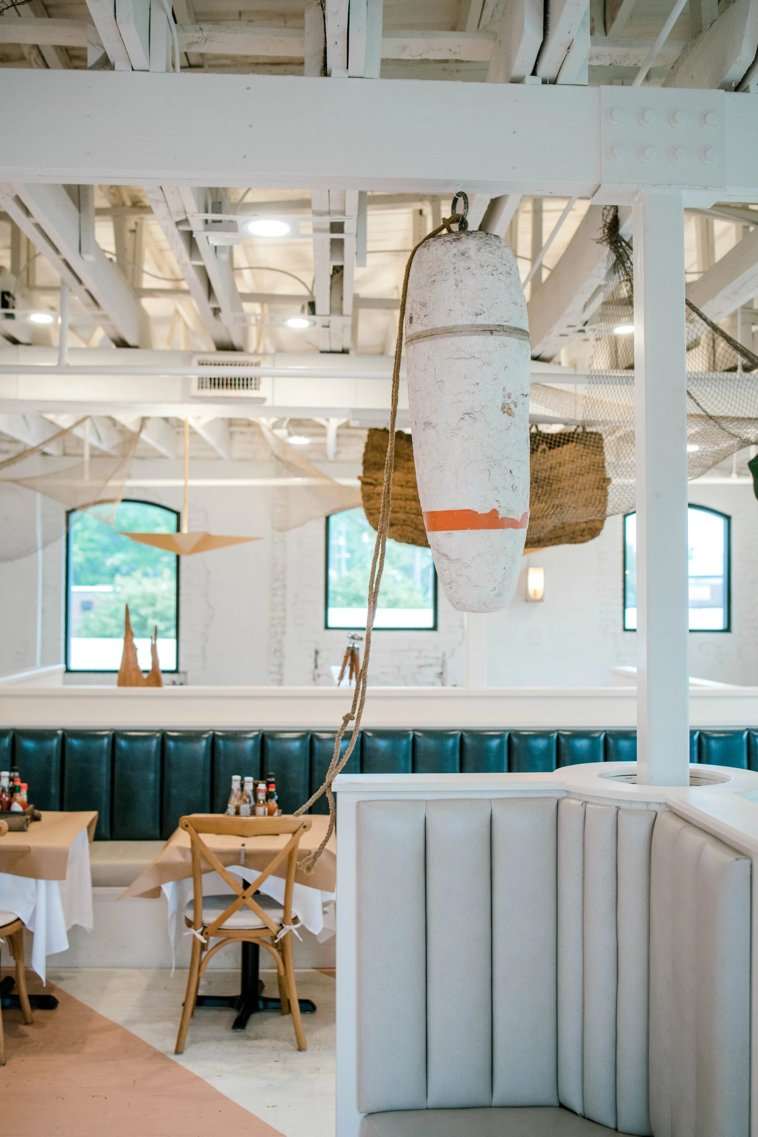 Interior of a restaurant with a buoys hanging from the ceiling, surrounded by white beams, wooden furniture, and large windows.