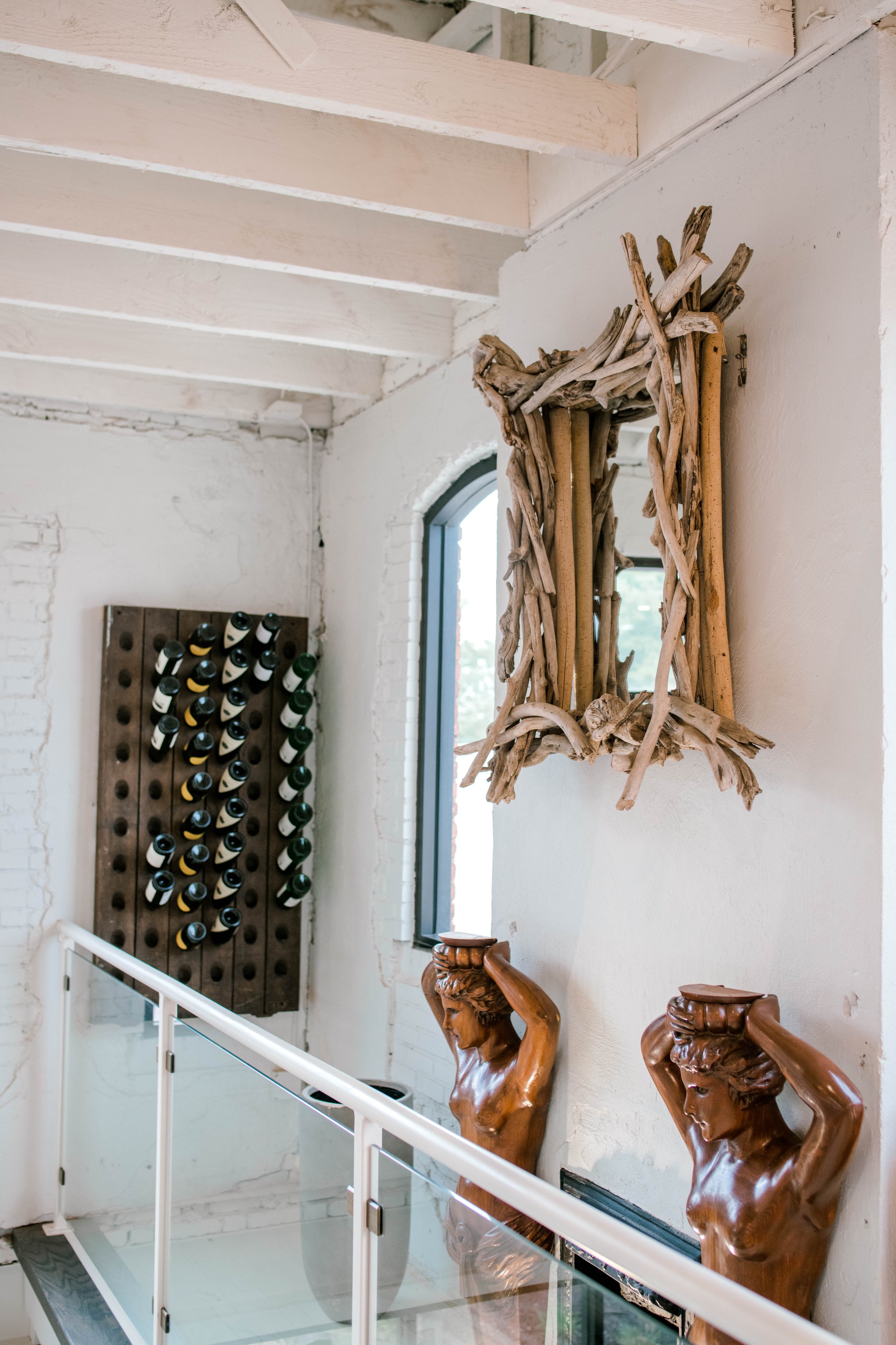 Interior of a building with a white brick wall, wooden sculpture of a woman, a mirror decorated with driftwood, a wine wine rack, and a glass railing.