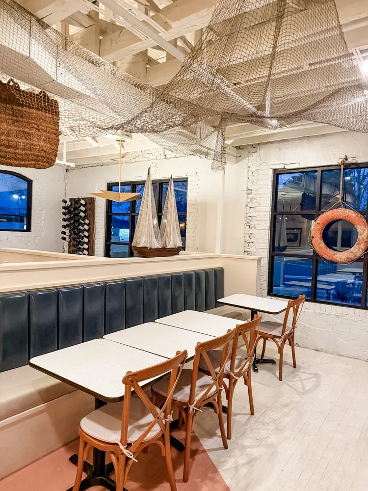 A cozy indoor dining area with white brick walls, black window frames, and nautical decor including a boat model, a lifebuoy, and fishing nets hanging from the ceiling. There is a long bench with dark upholstery and four wooden chairs in front of four white tables.