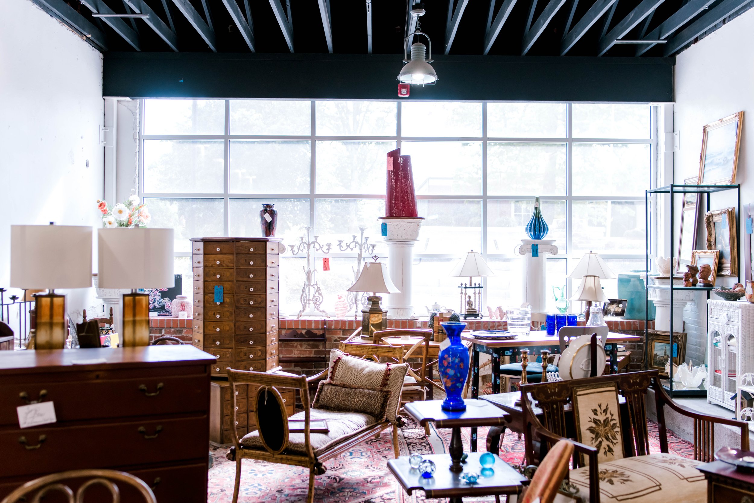 Interior of a thrift store or antique shop with various furniture and decorative items displayed, large window in the background, and industrial ceiling with hanging lights.
