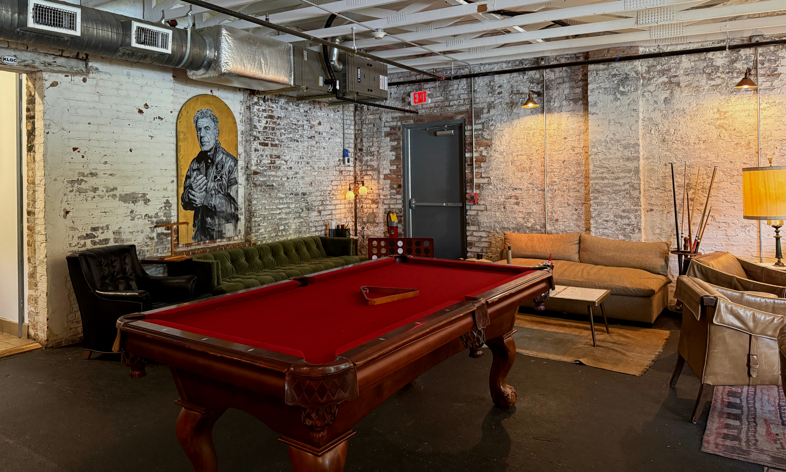 Interior of a cozy lounge or game room with exposed brick walls, featuring a red pool table in the center, a black leather armchair, a green tufted sofa, a beige sofa, various small tables, and wall-mounted lighting, with artwork on the wall and industrial ceiling elements.