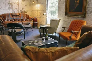 Cozy living room with vintage furniture including a brown leather sofa, an orange armchair, and a beige armchair. A dark coffee table holds magazines and candles. Exposed brick walls and large window with greenery outside are visible.