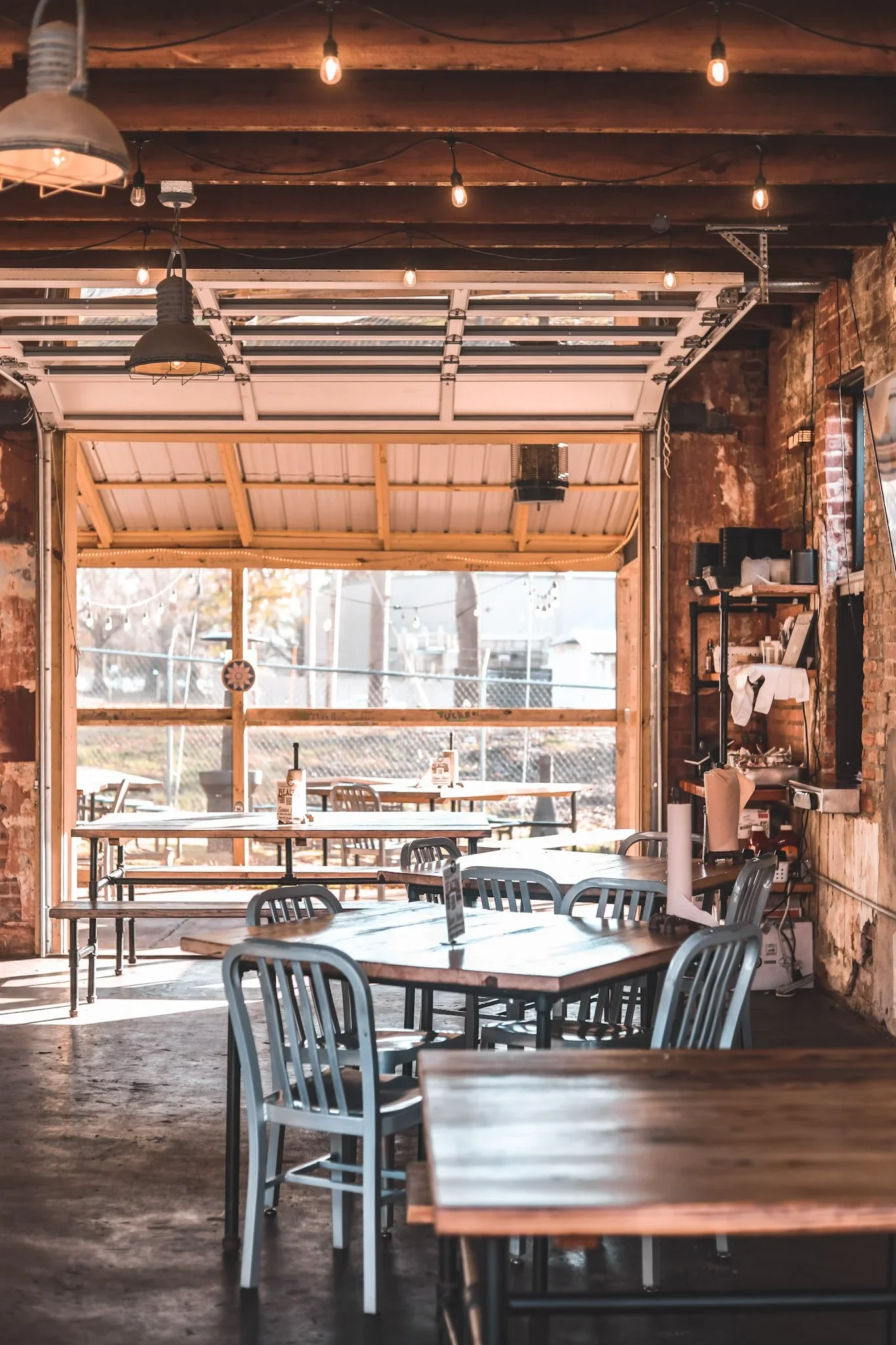 Interior of a rustic restaurant or cafe with wooden furniture and exposed brick walls, illuminated by Edison bulbs hanging from the ceiling. There are several tables and chairs, and large open garage-style doors allowing natural light to fill the space.