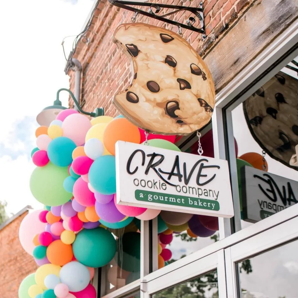 Colorful balloons adorn the storefront of CrAVE cookie company, a gourmet bakery, with a large cookie-shaped sign hanging above.
