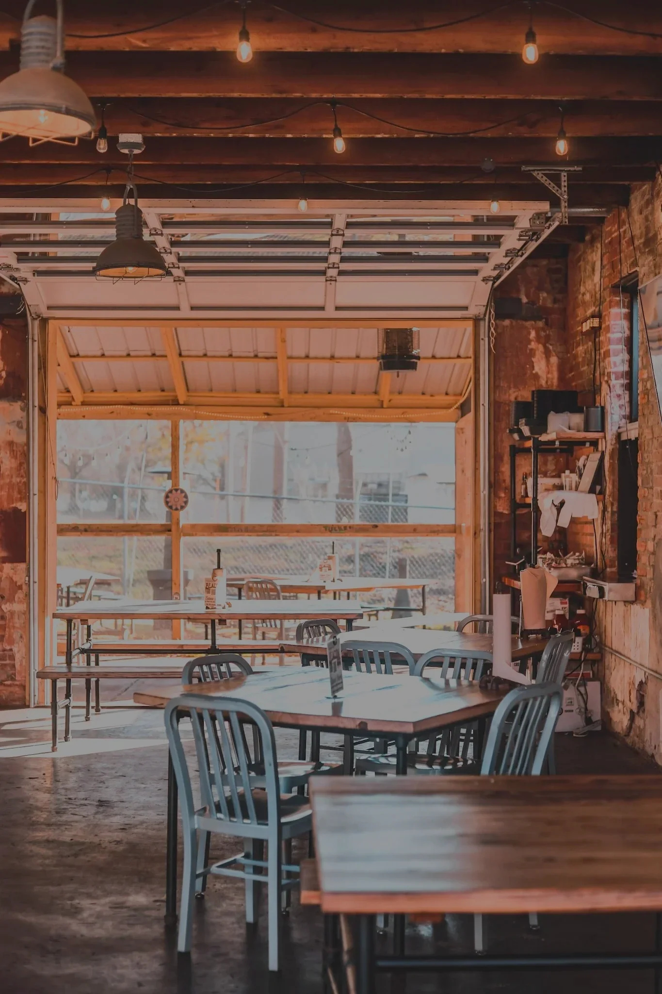 A cozy indoor dining area with wooden tables and blue chairs. Large windows allow natural light, and string lights are hanging from the ceiling.