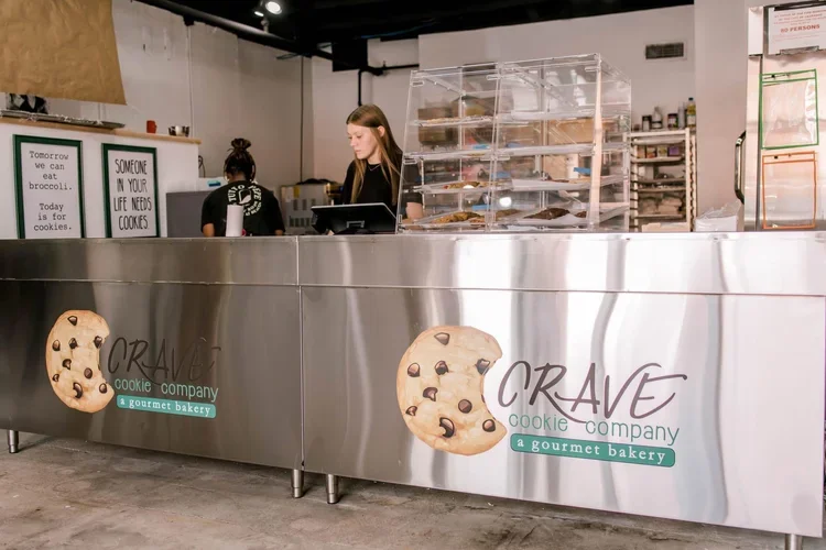 Front view of a bakery counter with silver metallic surface displaying the logo 'CRAVE cookie company' and images of cookies. Two women are working behind the counter with shelves of baked goods in the background.