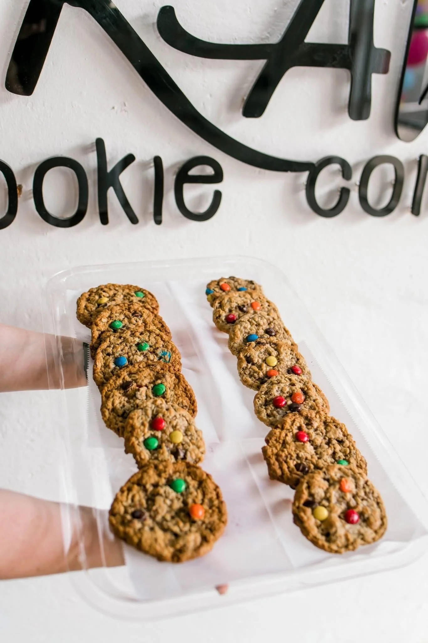 Close-up of chocolate chip cookies with colorful candies on a transparent tray against a white wall with black metal sign in the background.