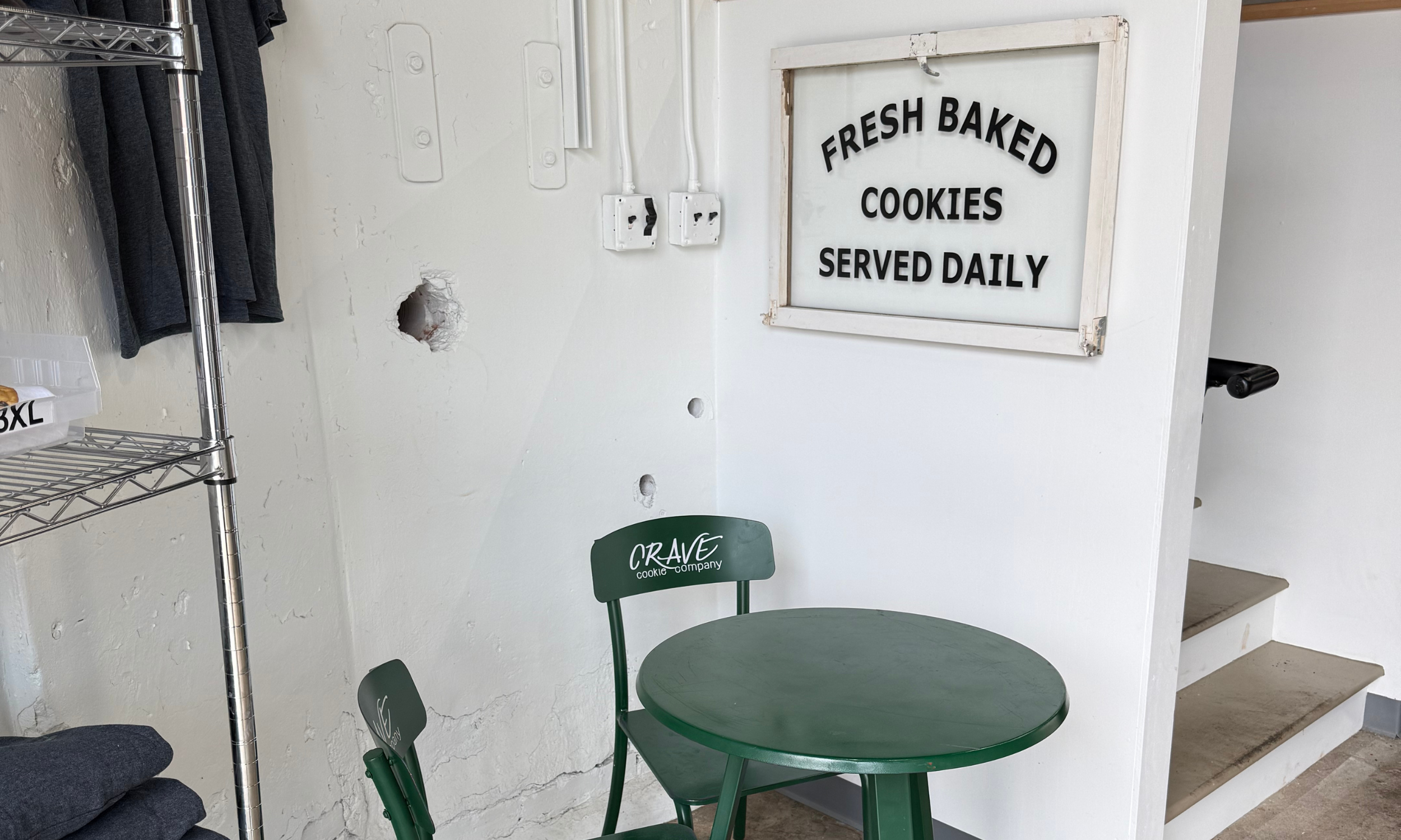 A corner of a room with a green round outdoor table and matching chairs, a white wall with power outlets, and a white sign with black text that reads "FRESH Baked Cookies Served Daily." There is a metal shelving unit with dark clothing on the left and some holes and patches on the wall.