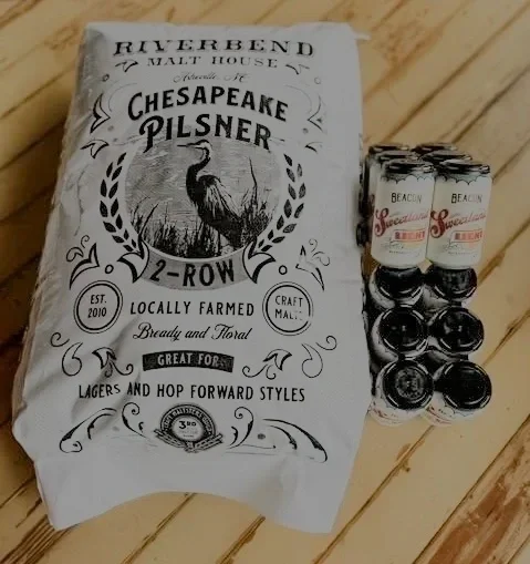 A bag of Chesapeake Pilsner 2-Row malt from Rivertend Malt House and several cans of Beech Nut Baby Food on a wooden surface.