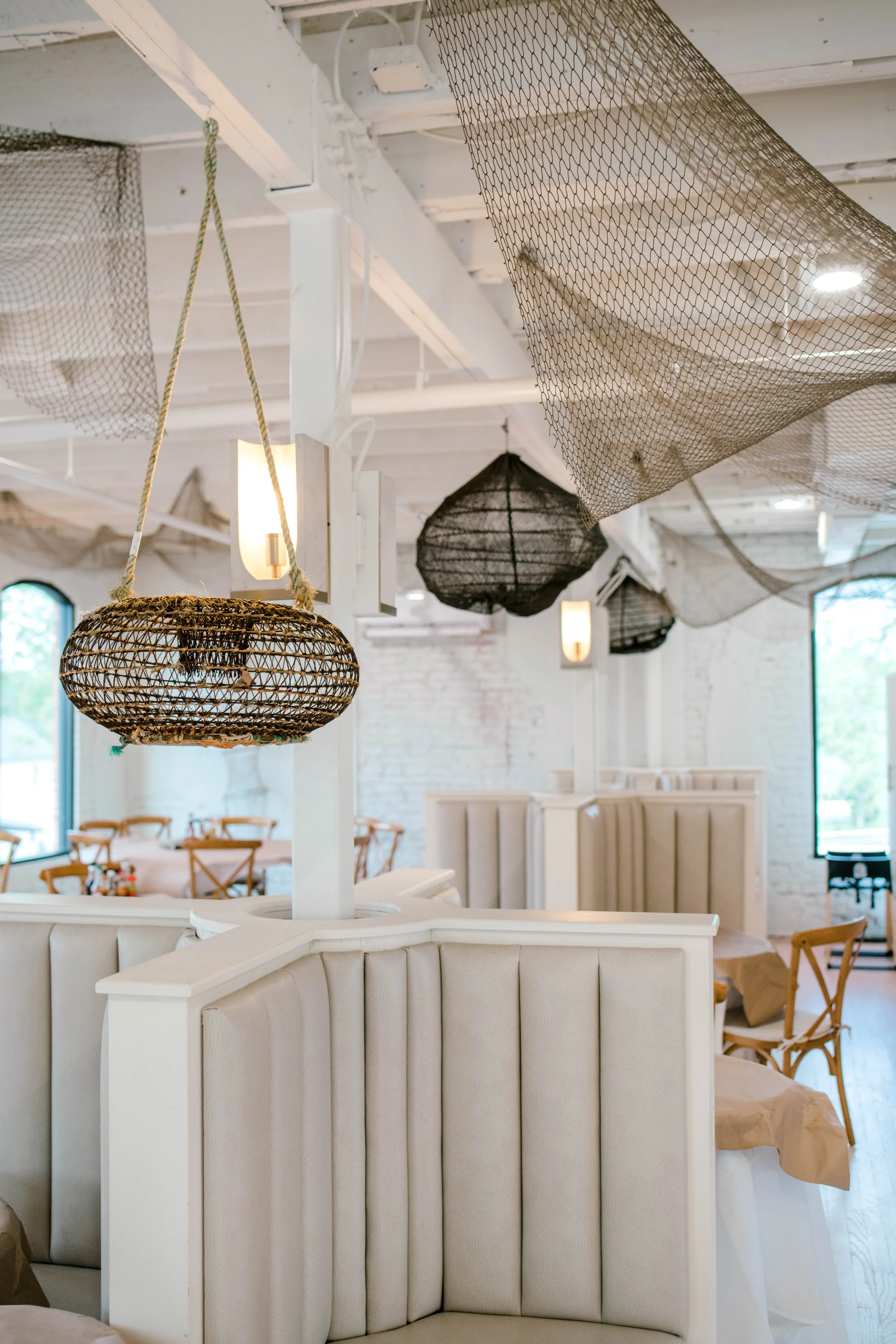 Interior of a restaurant with white walls and ceiling, decorated with hanging wicker lamps and black netting accents, wooden chairs, and white upholstered booths.