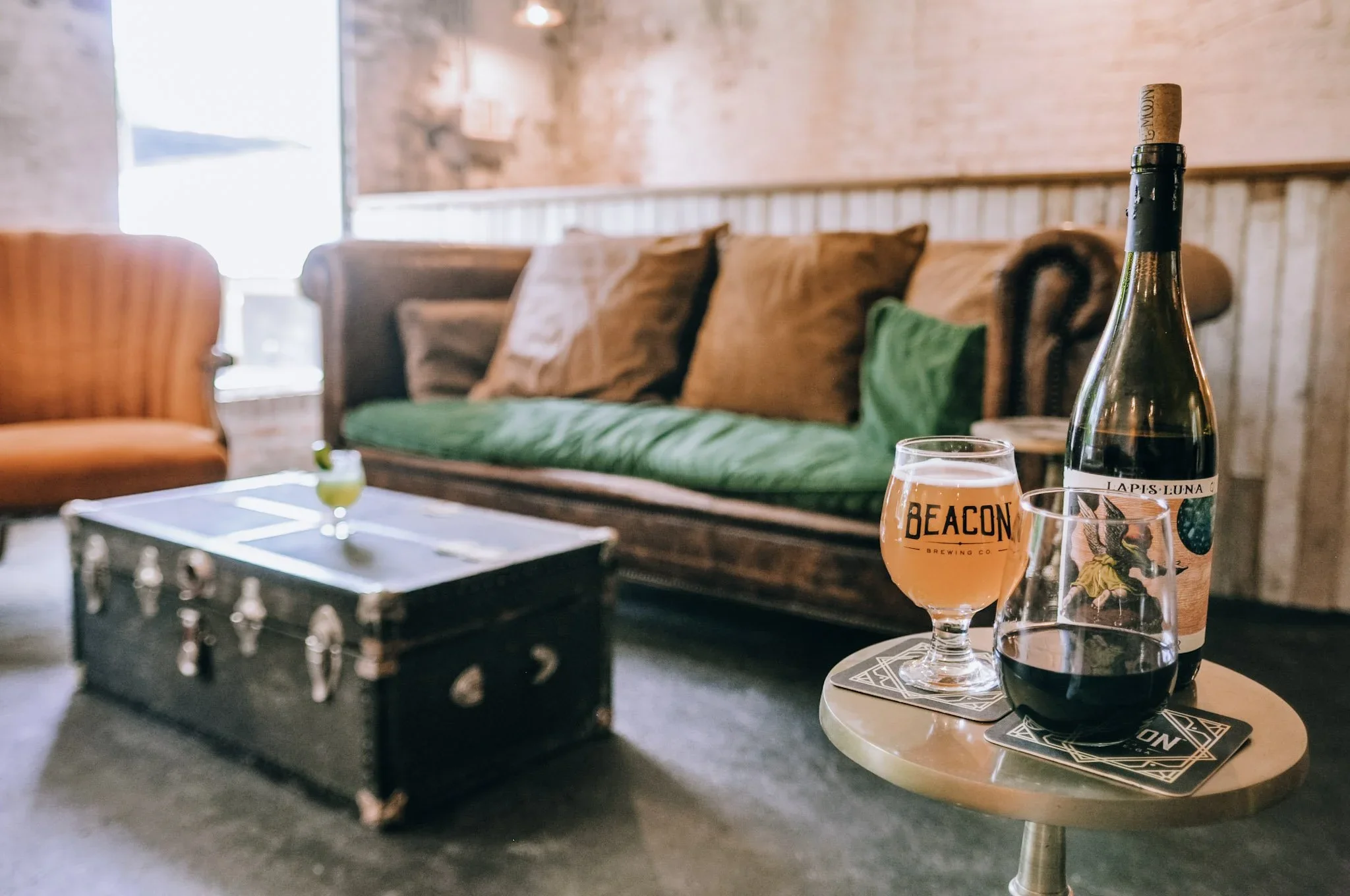 A cozy indoor space with a brick wall, brown and green cushions on a vintage sofa, a window allowing natural light, a small round table with two glasses of beer, a bottle of wine, and a foosball table in the foreground.