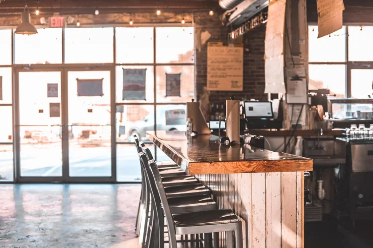 Inside a restaurant or bar with a long wooden counter, bar stools, and large windows letting in natural light. There are computer screens and menu boards behind the counter.