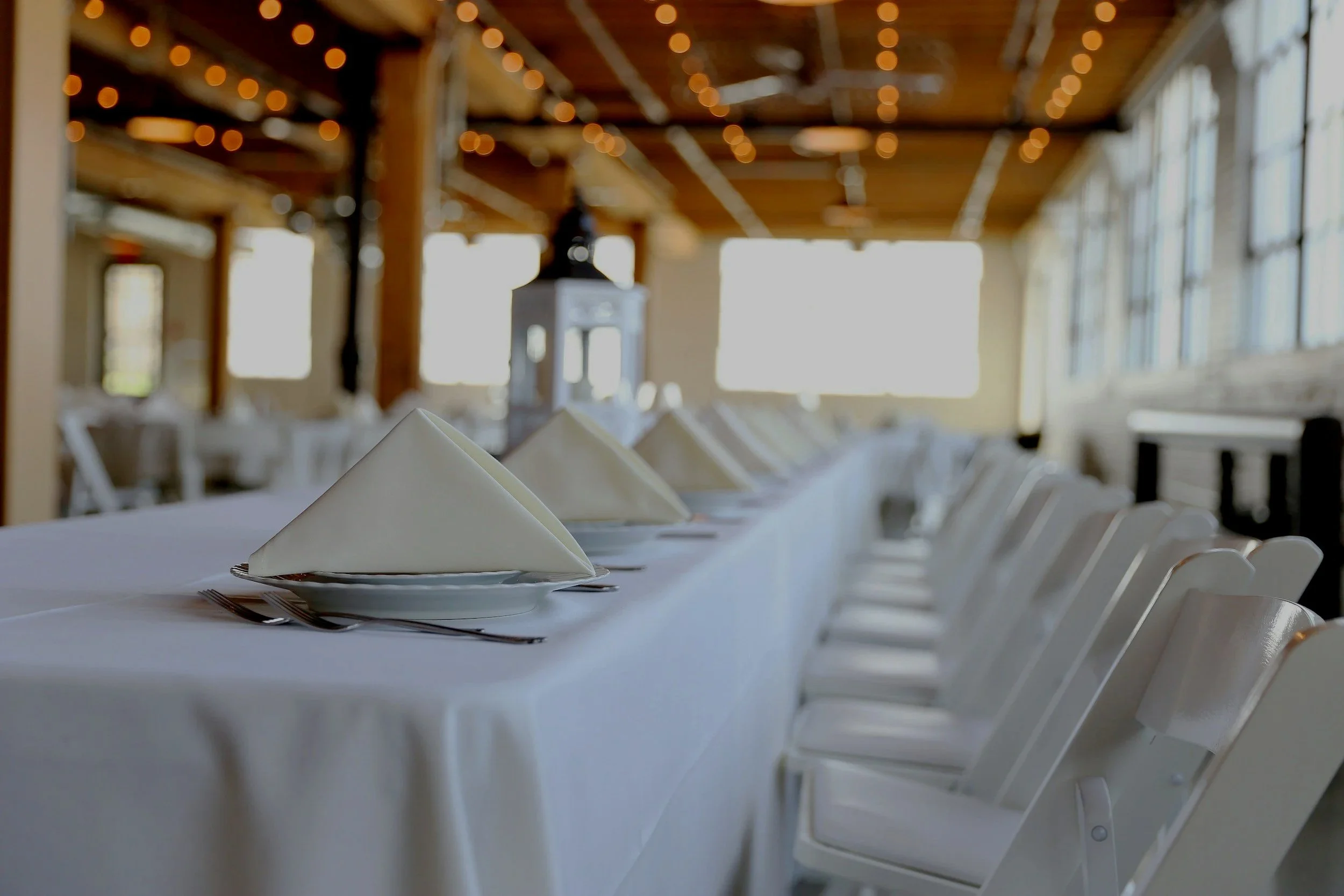 A long banquet table set for a meal with white tablecloths, white folded napkins, and silverware, in a bright room with large windows and string lights on a wooden ceiling.