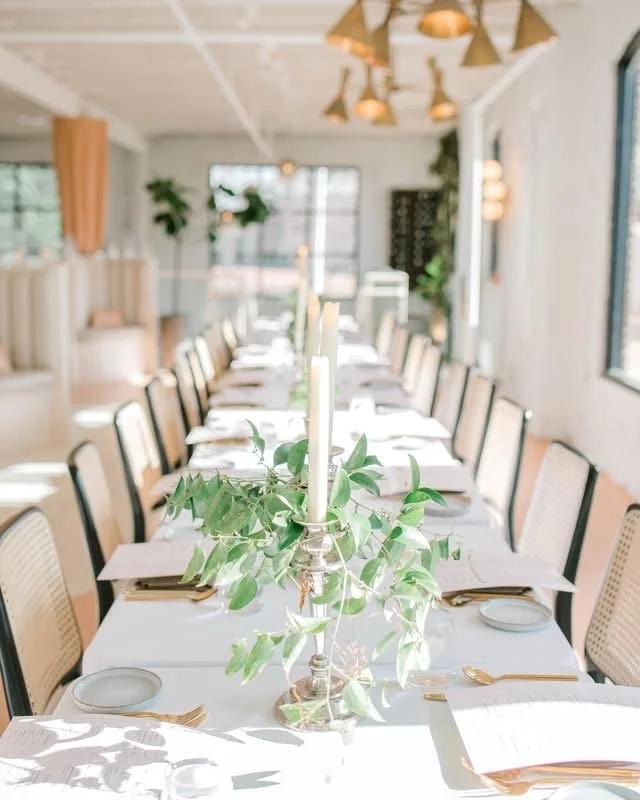 A long dining table set for a meal with white tablecloth, gold cutlery, white plates, and napkins, decorated with a central greenery arrangement and candles, in a bright, modern room with large windows and stylish lighting fixtures.