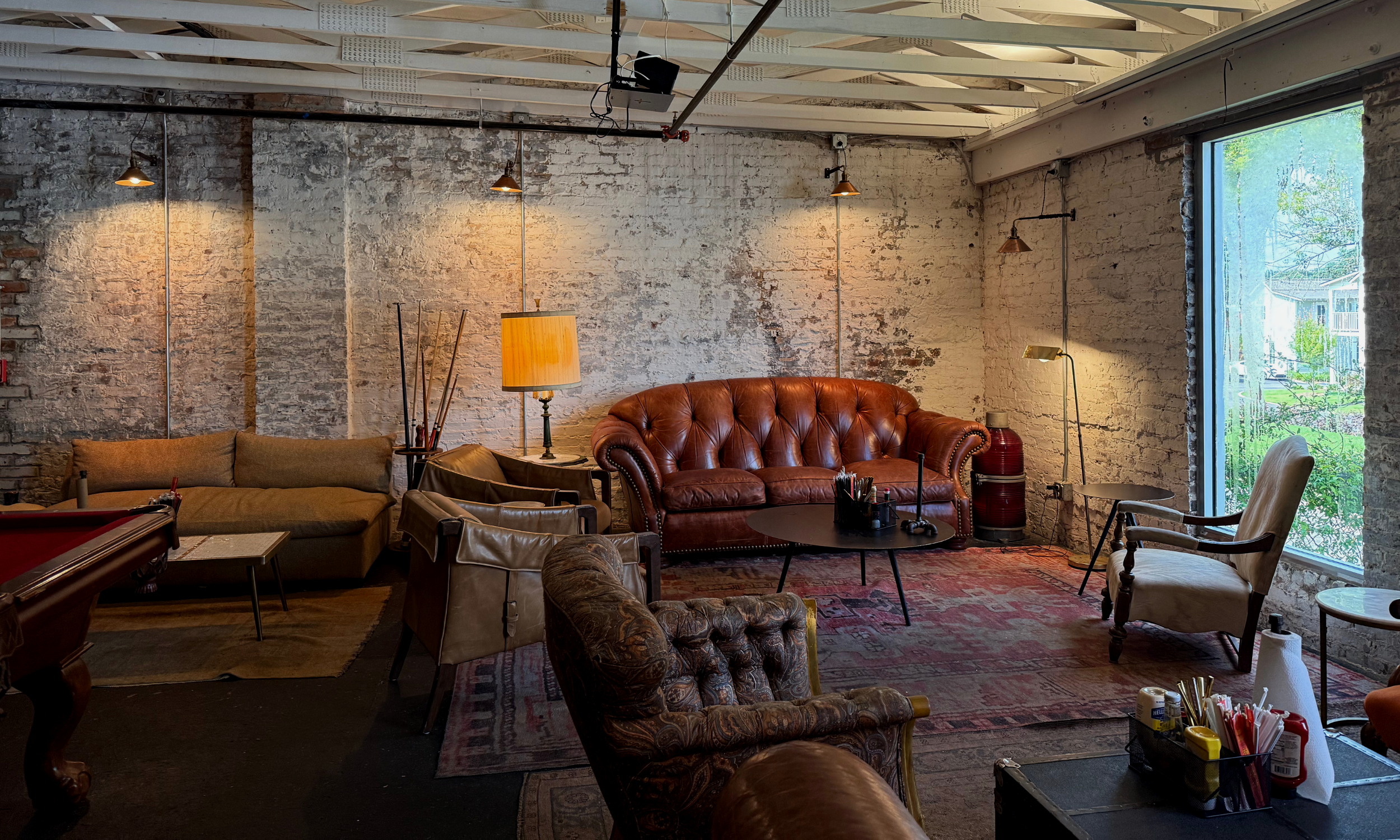 Cozy lounge area with exposed brick walls, vintage leather and fabric furniture, and various lamps, illuminated by natural light coming through a large window, suggesting an industrial-style interior.