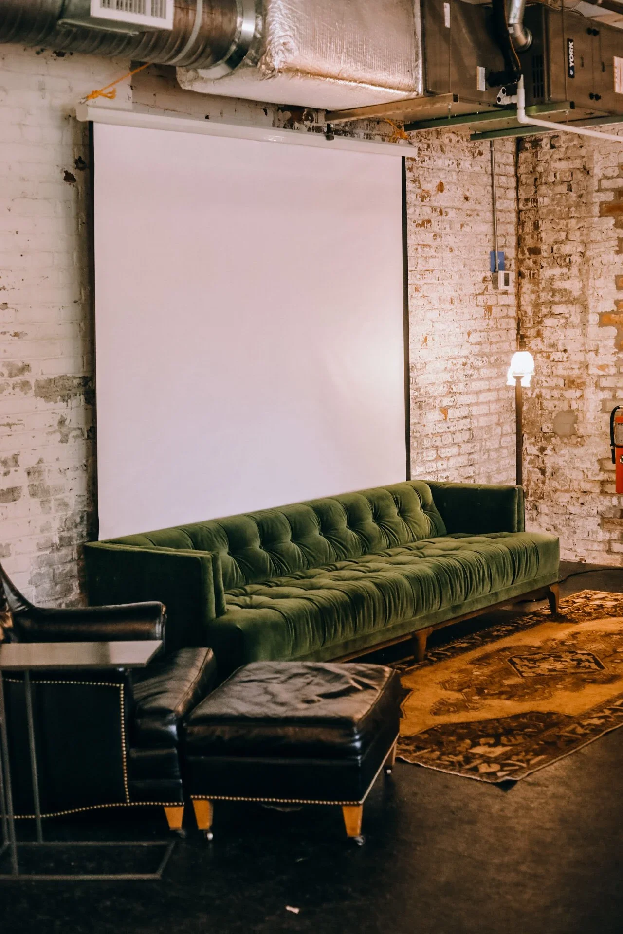A room with exposed brick walls, featuring a large white projection screen, a green velvet sofa, a black leather chair with a matching ottoman, a wooden floor with an area rug, and industrial lighting fixtures.