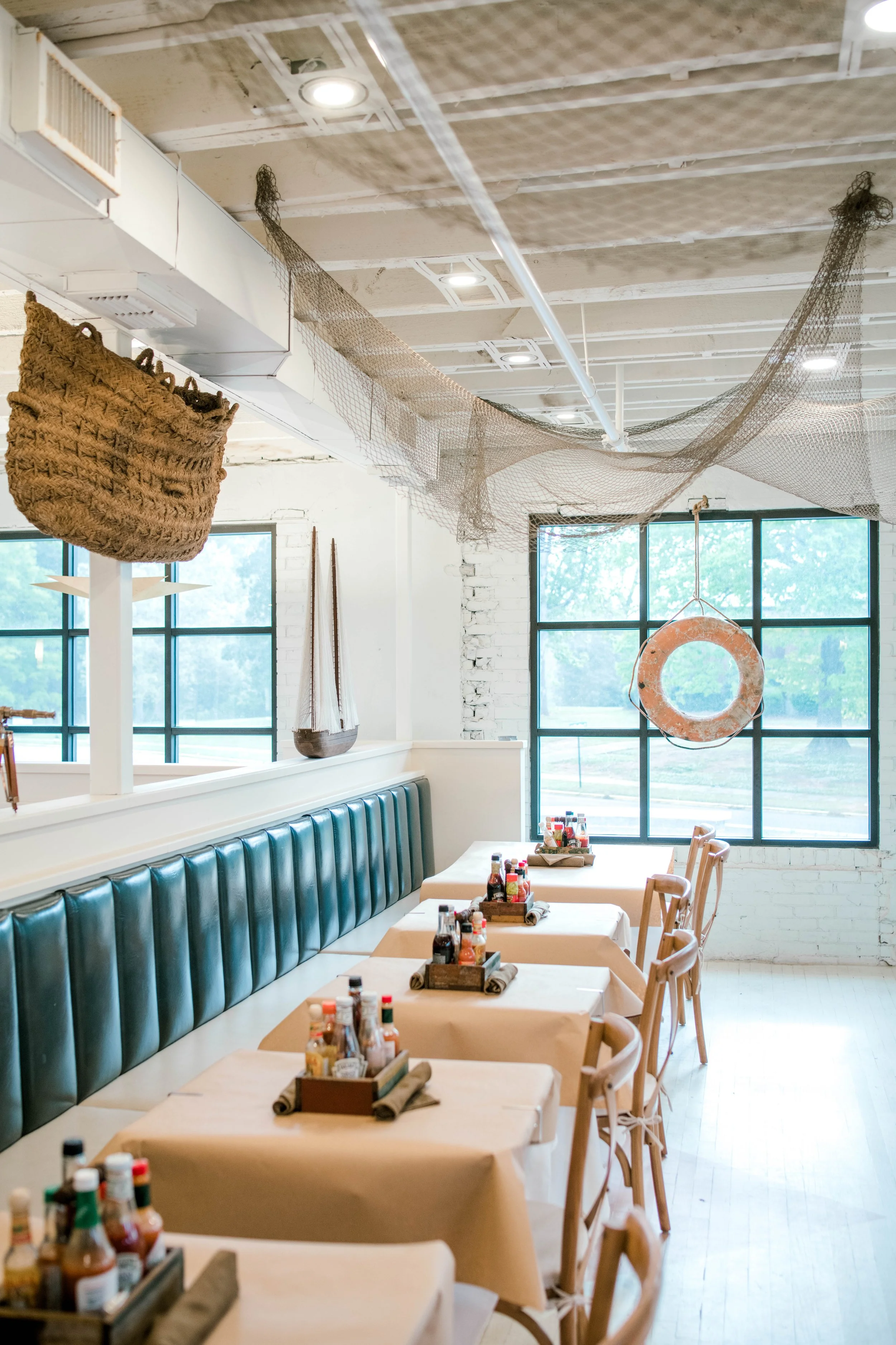 Interior of a restaurant with tables covered in beige tablecloths, wooden chairs, and teal upholstered bench seating. The tables are set with condiment trays, and the room features large windows with a view of trees outside. The ceiling has decorative fishing nets and natural light.