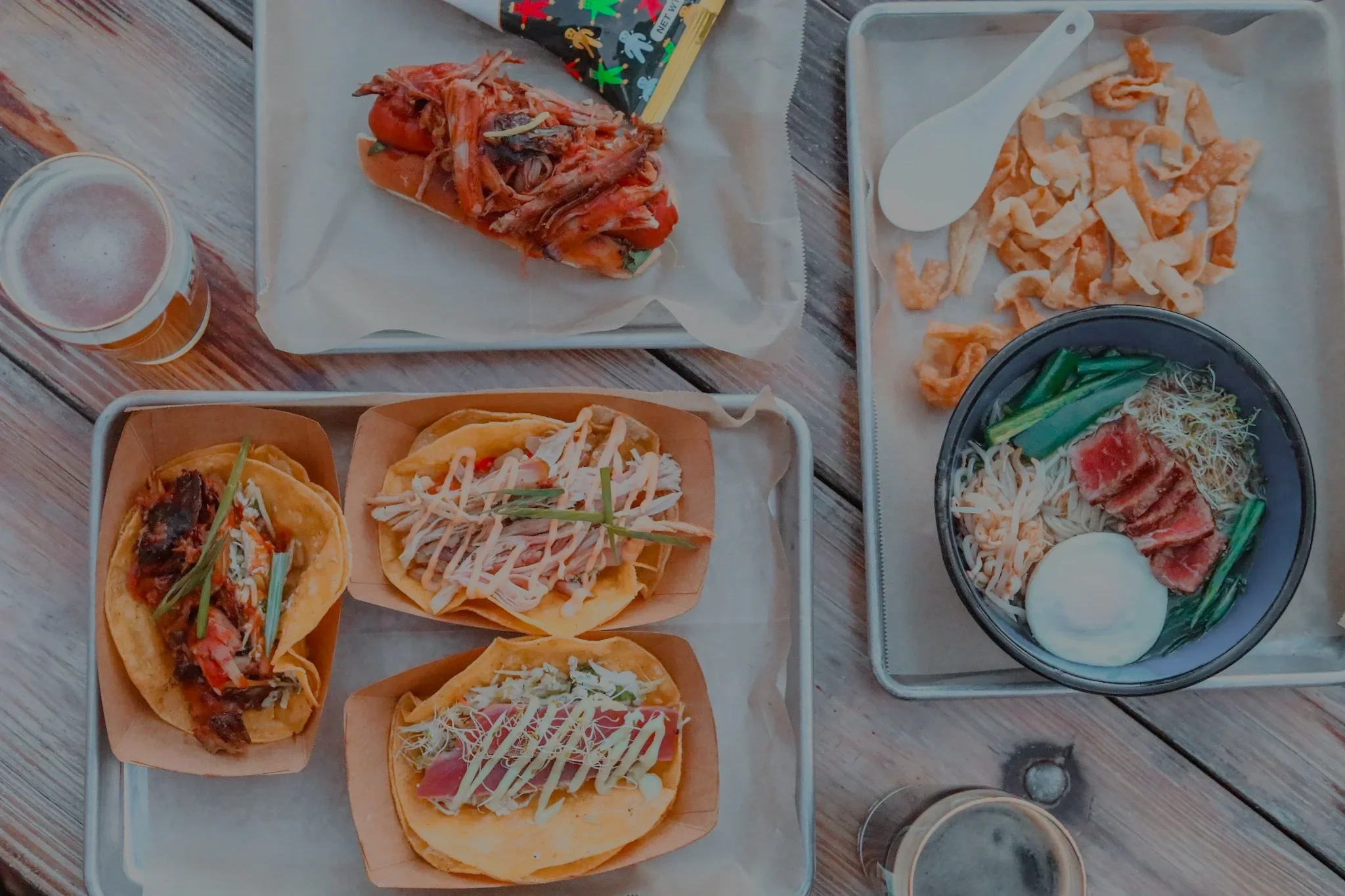 Various tacos with different fillings, a bowl of ramen, a plate of fried food, a pint of beer, and a tray of food on a wooden table.