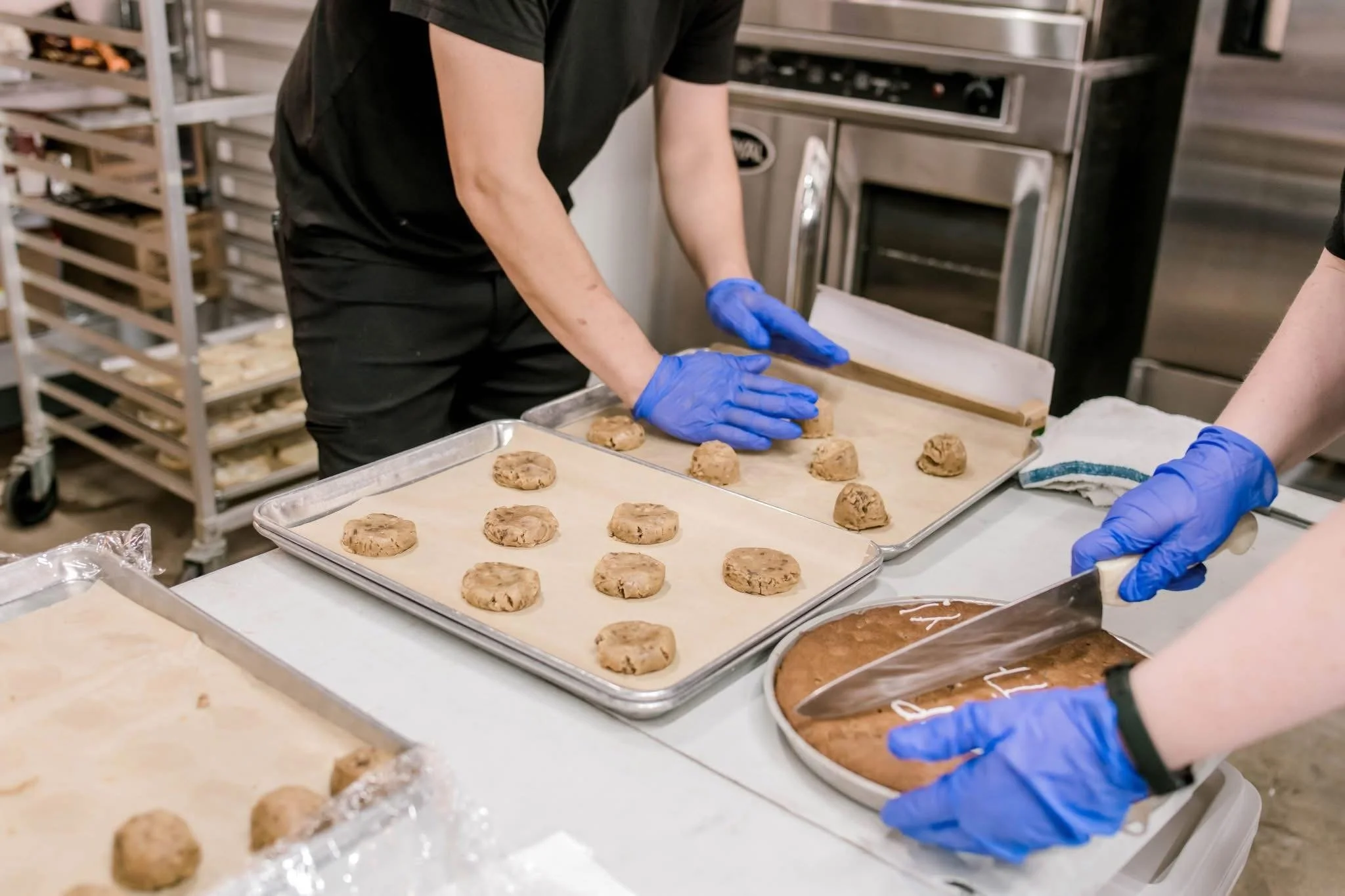 A person wearing black clothing and blue gloves is placing cookie dough balls on a baking sheet in a commercial kitchen.