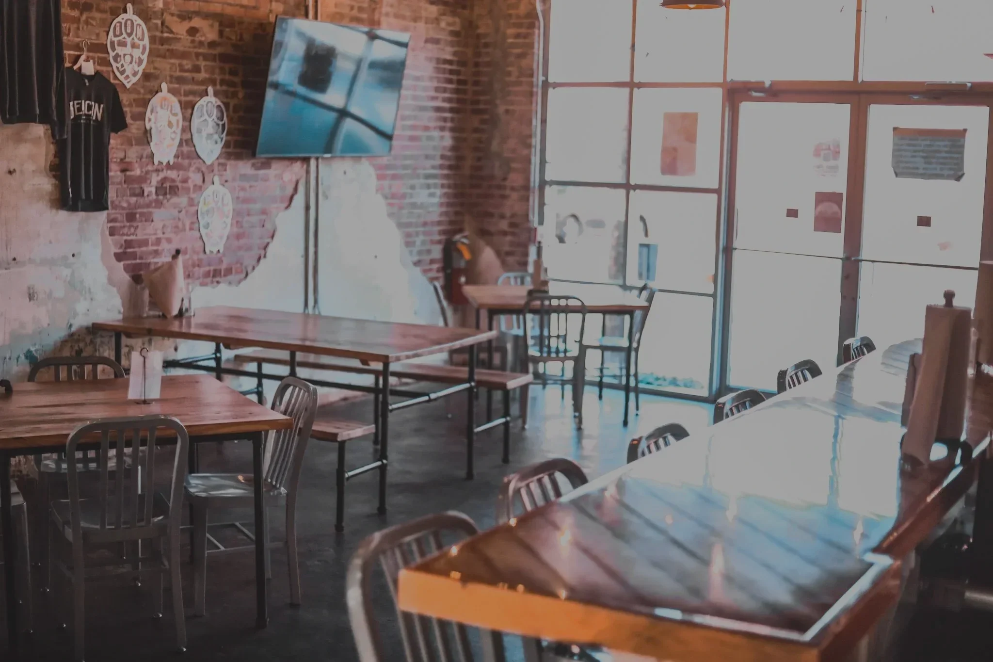 Inside a cafe or restaurant with wooden tables and metal chairs, brick walls, a wall-mounted TV, and large windows letting in natural light.