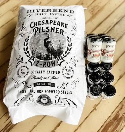 A bag of Riverbend Chesapeake Pilsner malt and a six-pack of cans labeled 'Beacon Special Lager' on a wooden surface.