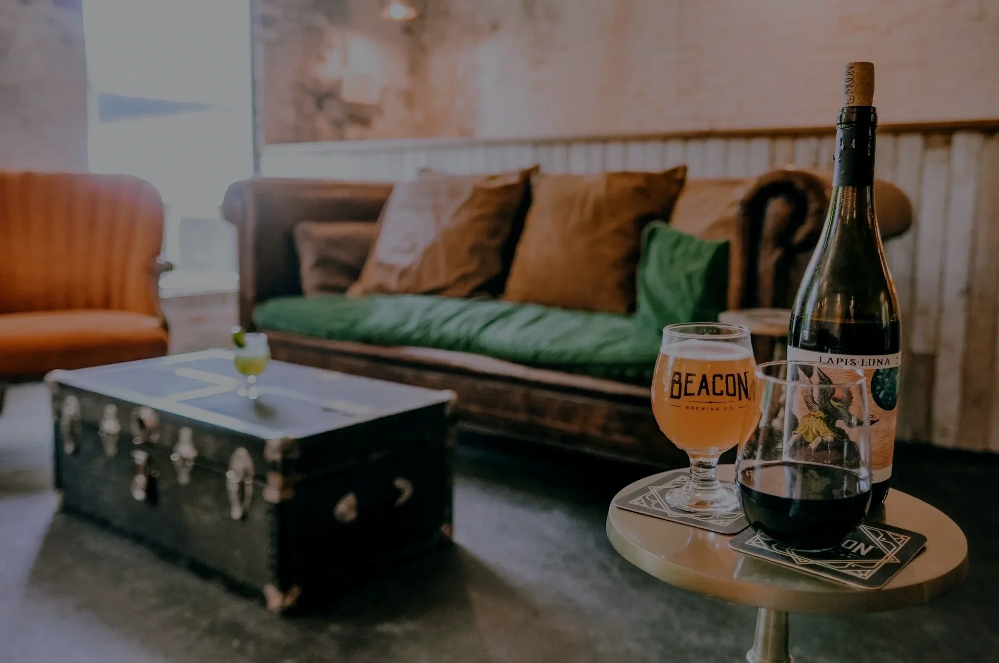 A bar setting with a bottle of wine and two glasses of beer on a small round table, with a green cushioned bench, brown pillows, and a wooden wall in the background.