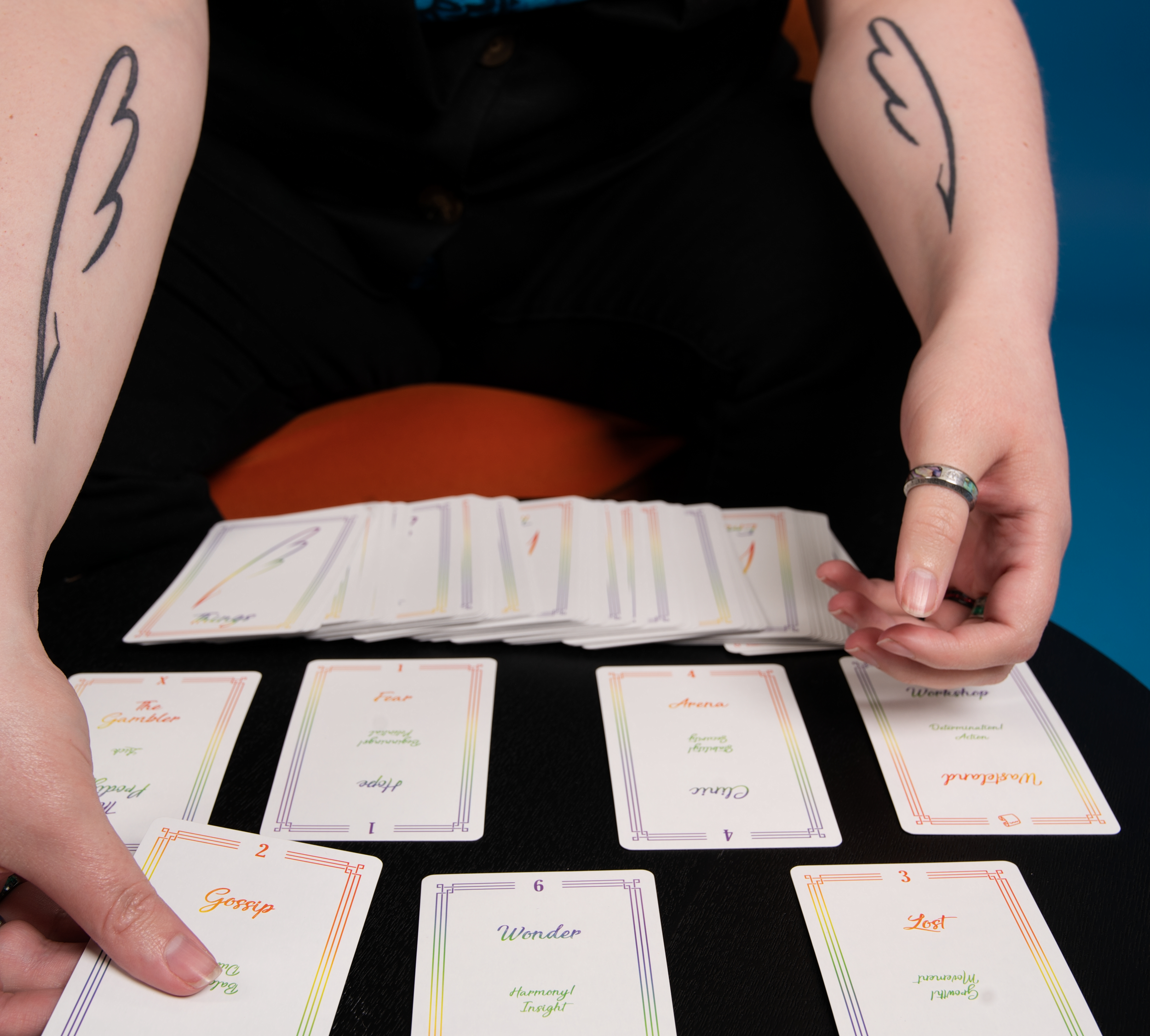 Person holding a fan of colorful cards with words like "Gossip," "Fame," and "Wonder," in front of a table with more cards and a tattooed arm in the background.