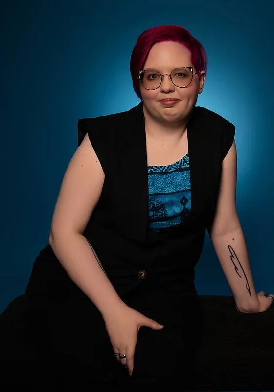 A person with short pink hair and glasses, wearing a black vest and blue patterned top, sitting against a blue background.