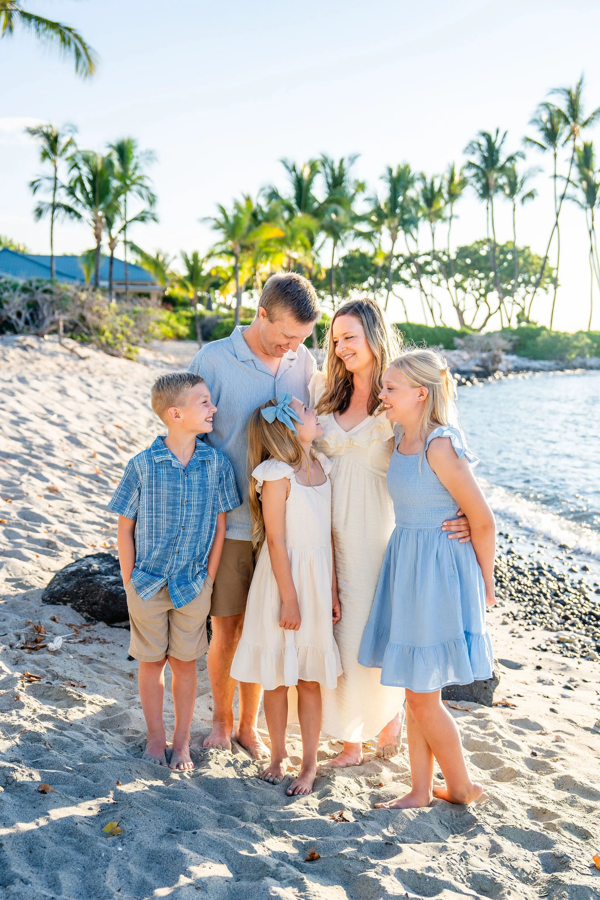 A family of six, including two adults and four children, standing on a sandy beach with palm trees in the background. They are smiling and looking at each other, enjoying a sunny day by the water.
