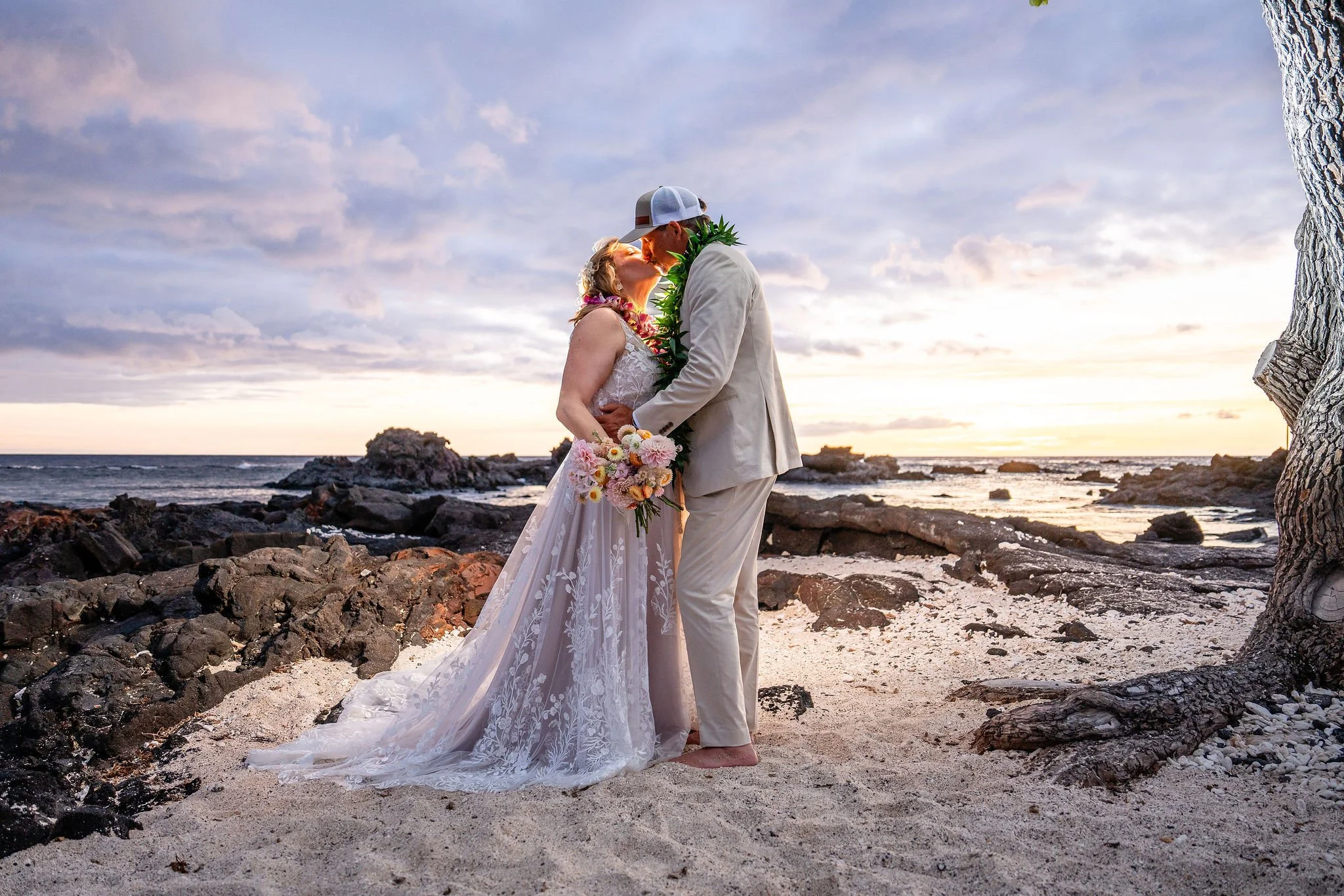 A couple getting married on a beach during sunset, with the bride in a white dress holding a colorful bouquet and the groom in a light suit and baseball cap, sharing a kiss.